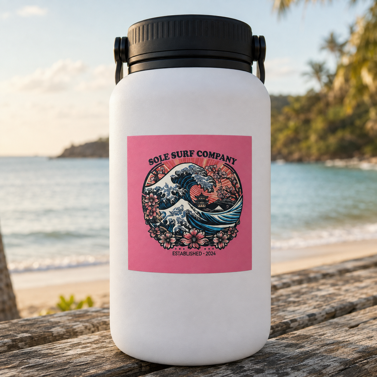 A white insulated water bottle with a black lid sits on a wooden table outdoors, with a beach and ocean blurred in the background. The bottle has a pink square sticker reading 'SOLE SURF COMPANY' with a wave and floral illustration.