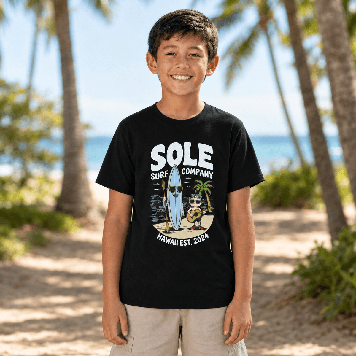 A smiling young boy stands on a sandy beach wearing a black graphic t-shirt with 'SOLE SURF COMPANY' and a cartoon surfboard character, set against a blurred background of palm trees and the ocean.
