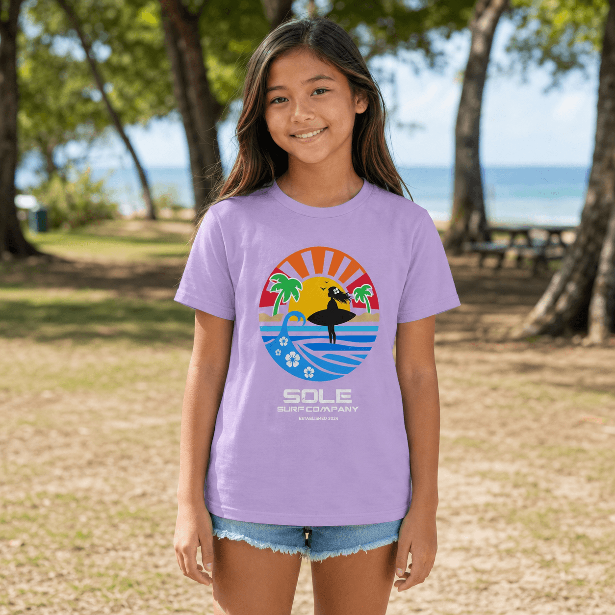 A young girl stands outdoors in a park-like setting near trees and the ocean, wearing a light purple graphic T-shirt with a retro surfboard-themed circular print and the text 'SOLE SURF COMPANY' on the front, paired with denim shorts. This is the girls surf series tee