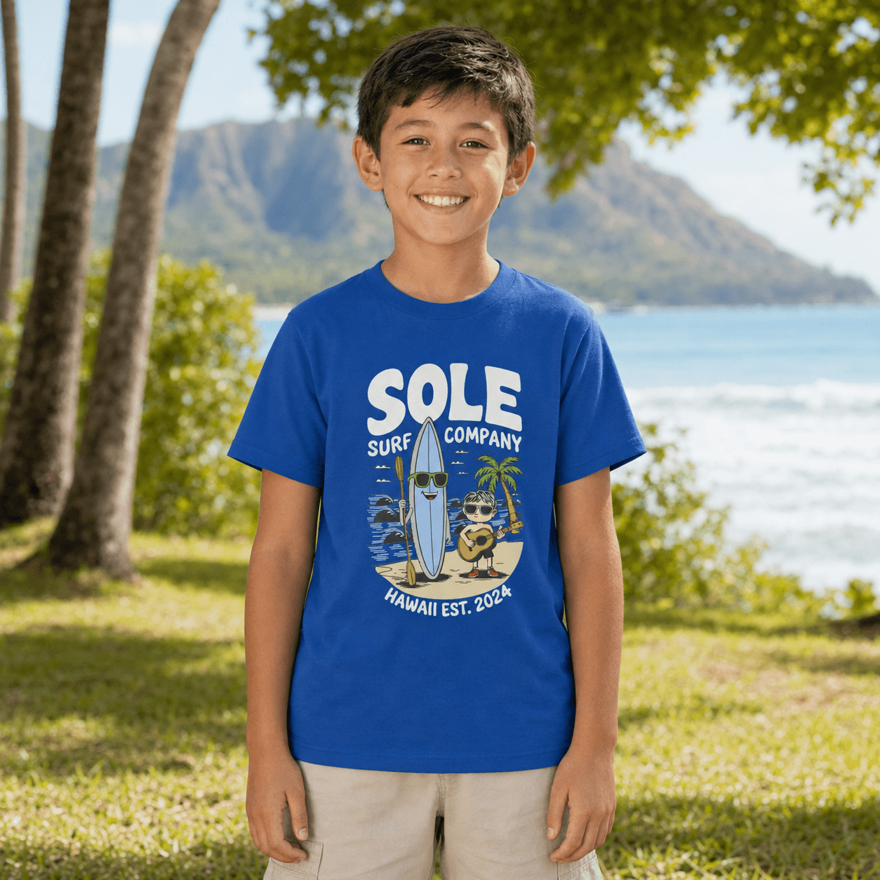 A smiling young boy stands outdoors in front of a tropical beach and mountains, wearing a bright royal blue graphic T-shirt with 'SOLE SURF COMPANY' and a surfboard illustration on the front, paired with light beige shorts.