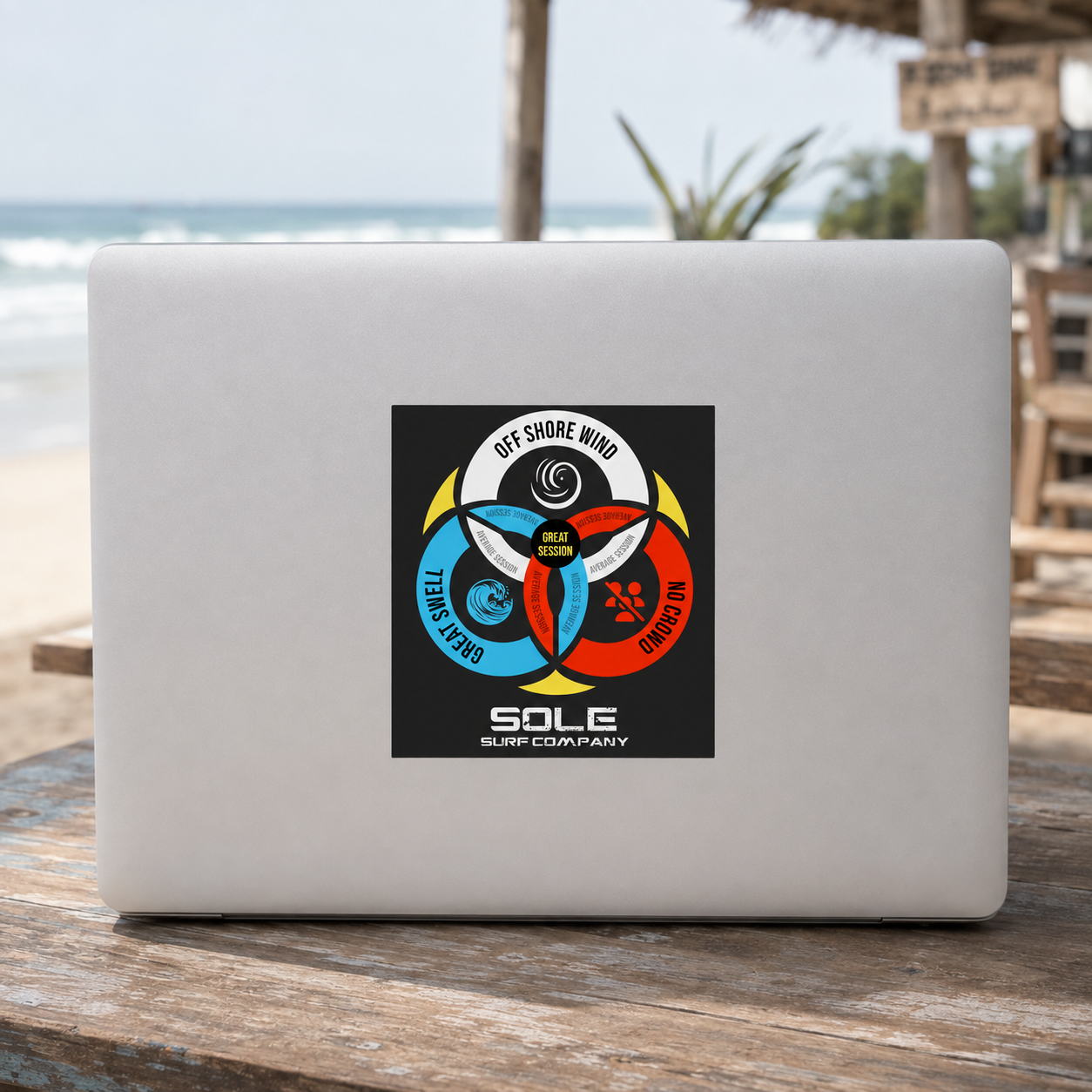 A silver laptop resting on a wooden table at a beach, featuring a colorful circular graphic sticker on the back. The sticker displays the text 'GET SHORE WIND' and 'SALE SURF COMPANY' with three colored segments (white, blue, red) and a central yellow circle.