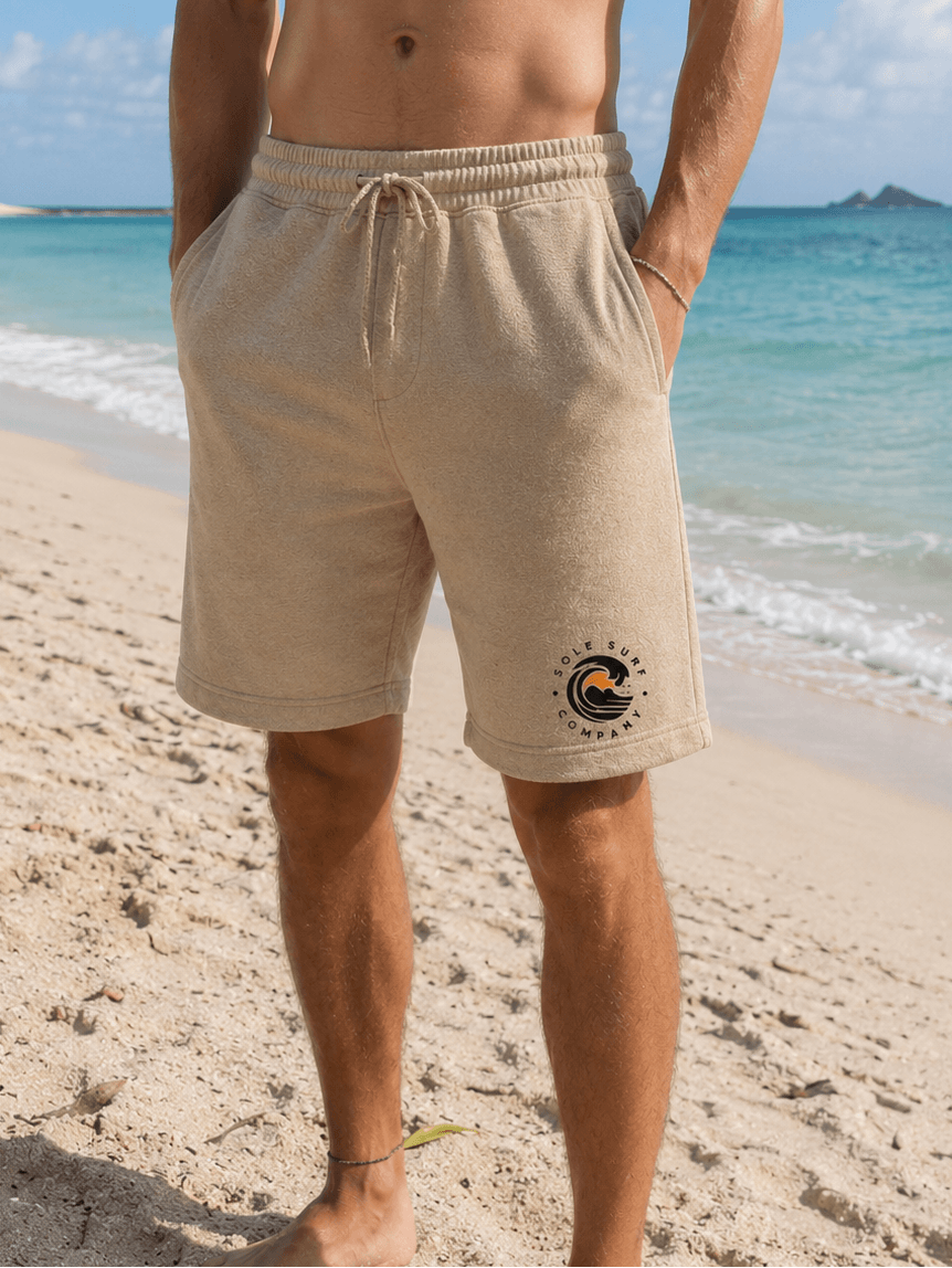 A man standing on a sandy beach wearing beige drawstring shorts with a small circular wave graphic on the lower left leg. The background shows turquoise ocean water and a blue sky.