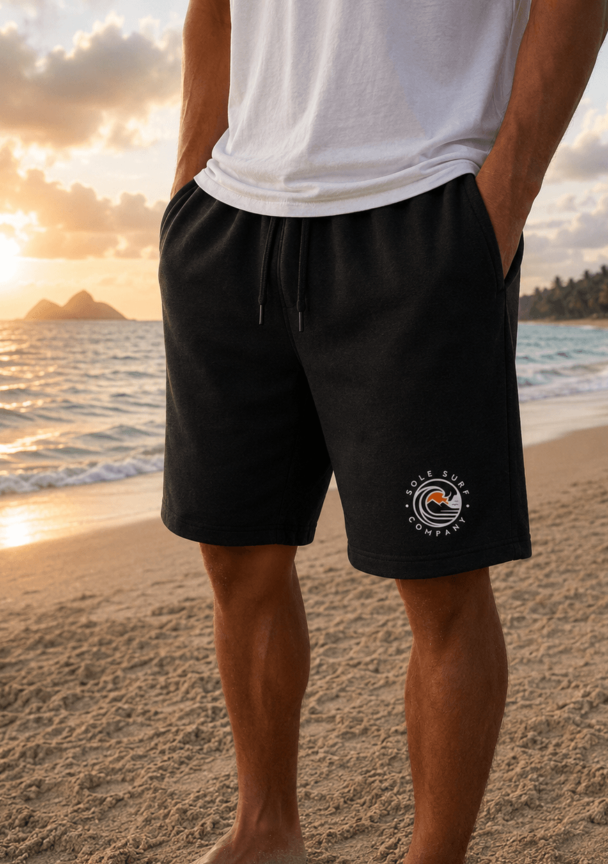 A person standing on a sandy beach at sunset wearing black drawstring shorts with a circular 'Colder Company' logo on the lower left leg, paired with a white t-shirt.