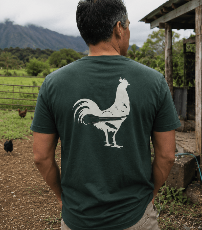 Man wearing a green t-shirt with a rooster design, standing in a farm setting with chickens and a mountain in the background.