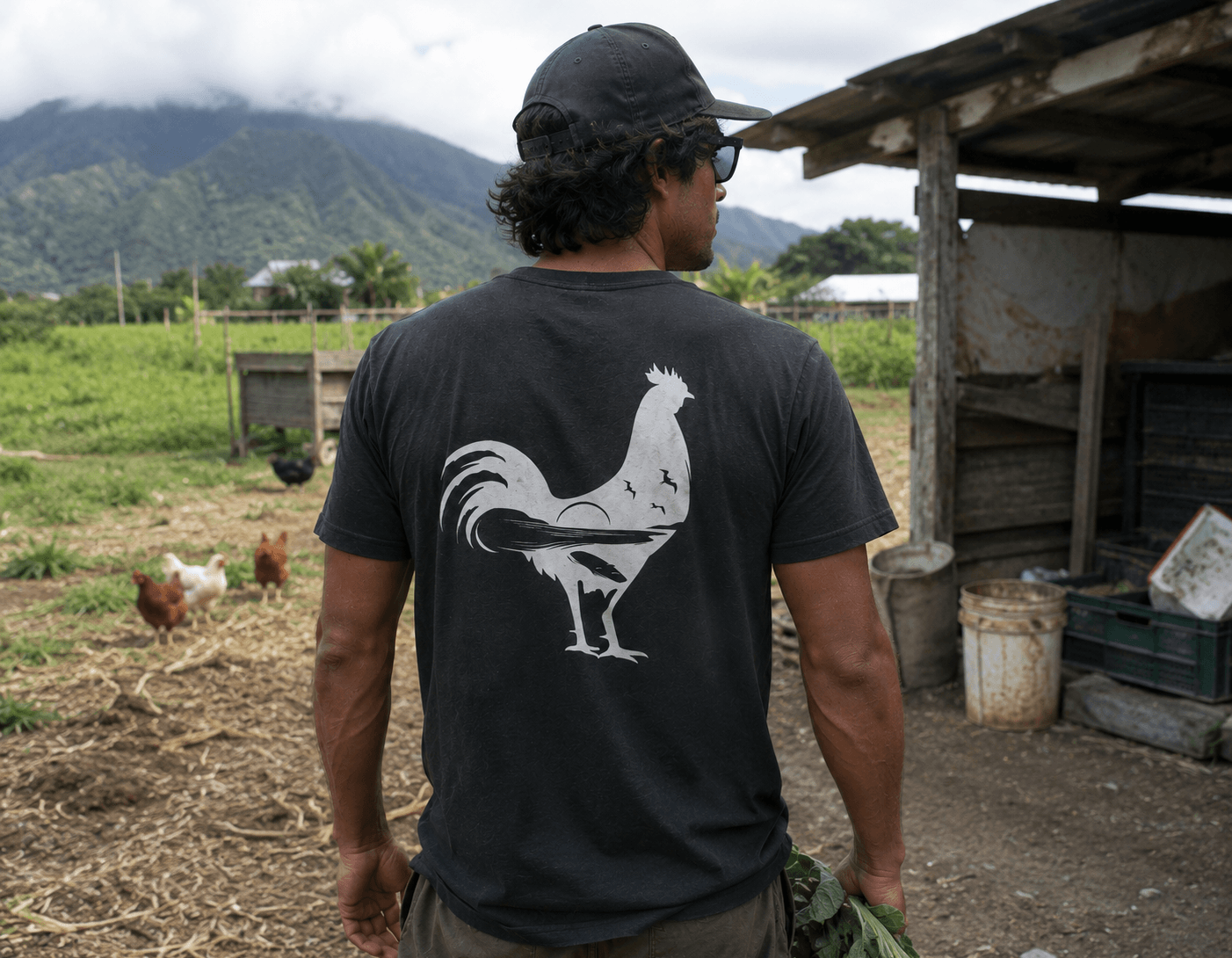 Man wearing a t-shirt with a rooster design on a farm with chickens and mountains in the background.
