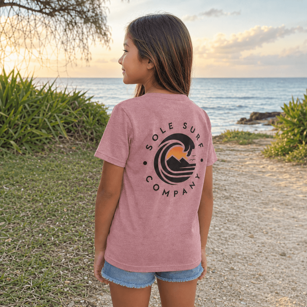 A young girl stands on a beach path at sunset wearing a heathered pink t-shirt with a large black and orange wave graphic and the text 'SOLE SURF COMPANY' on the back, paired with denim shorts.