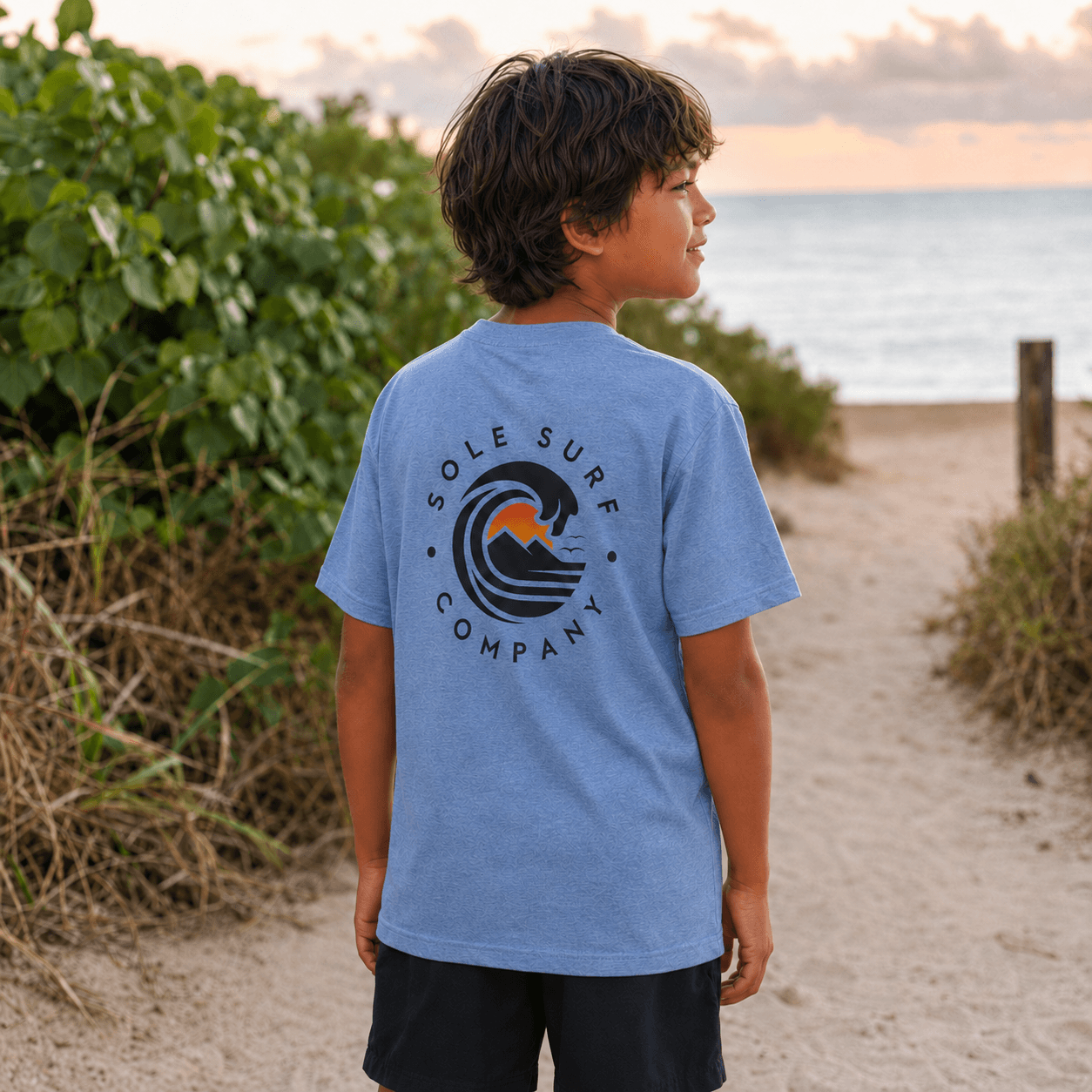 A child wearing a heathered light blue t-shirt with a circular 'Sole Surf Company' logo on the back, standing on a sandy beach path with green plants and the ocean in the background.
