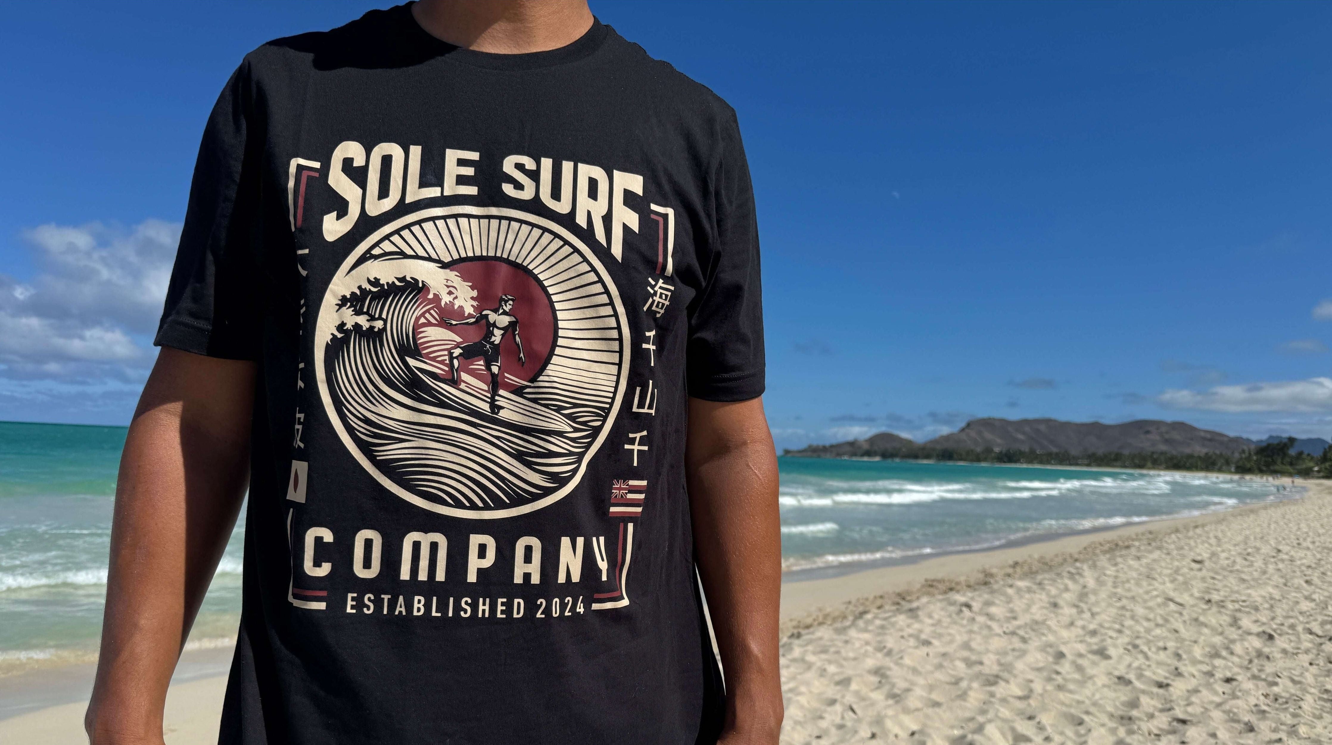 A person wearing a black graphic t-shirt with 'SOLE SURF COMPANY' printed on the front, standing on a sandy beach with the ocean and blue sky in the background.