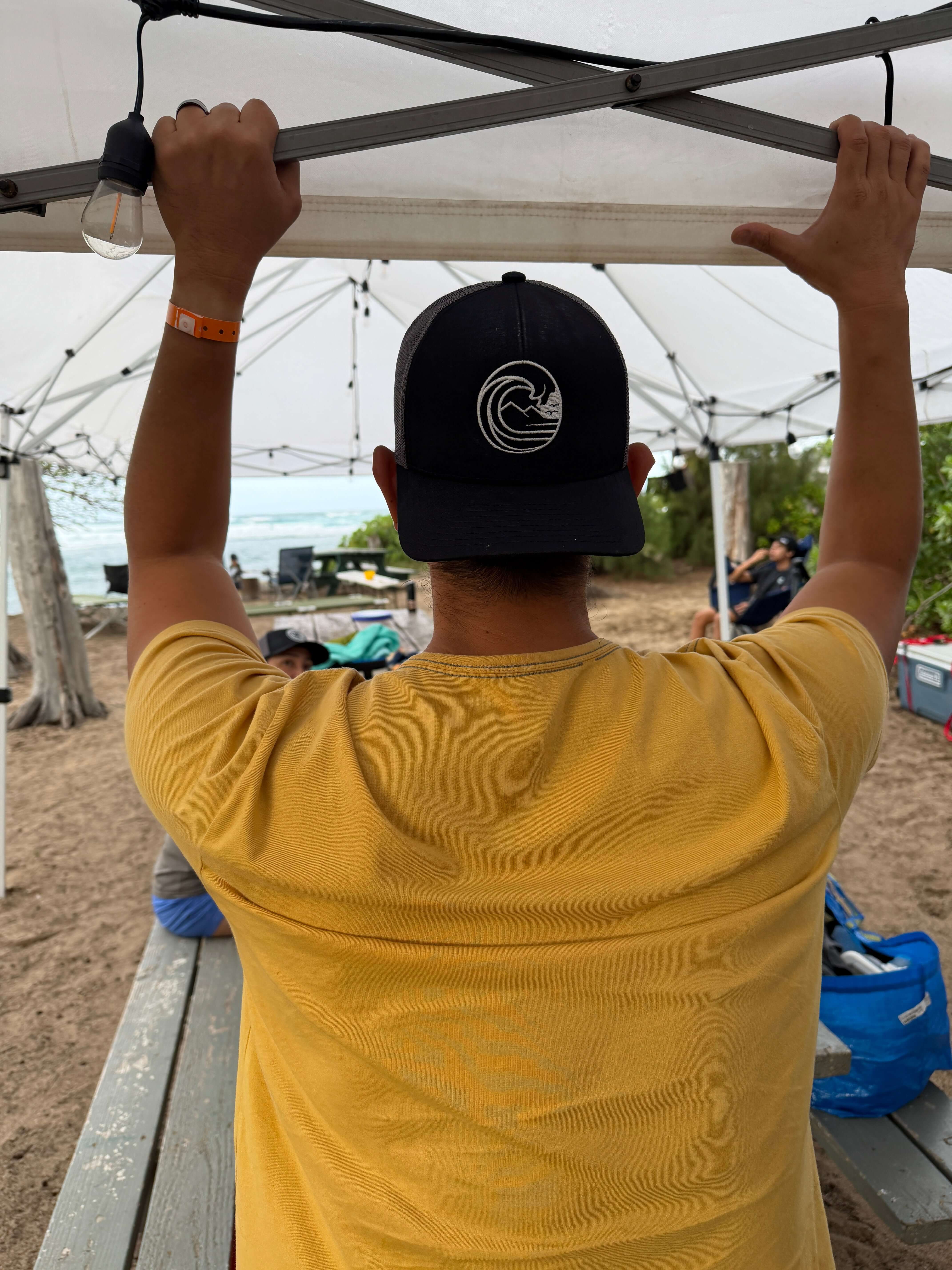 Person wearing SSC Trucker Cap, enjoying a beach day under a tent with friends.
