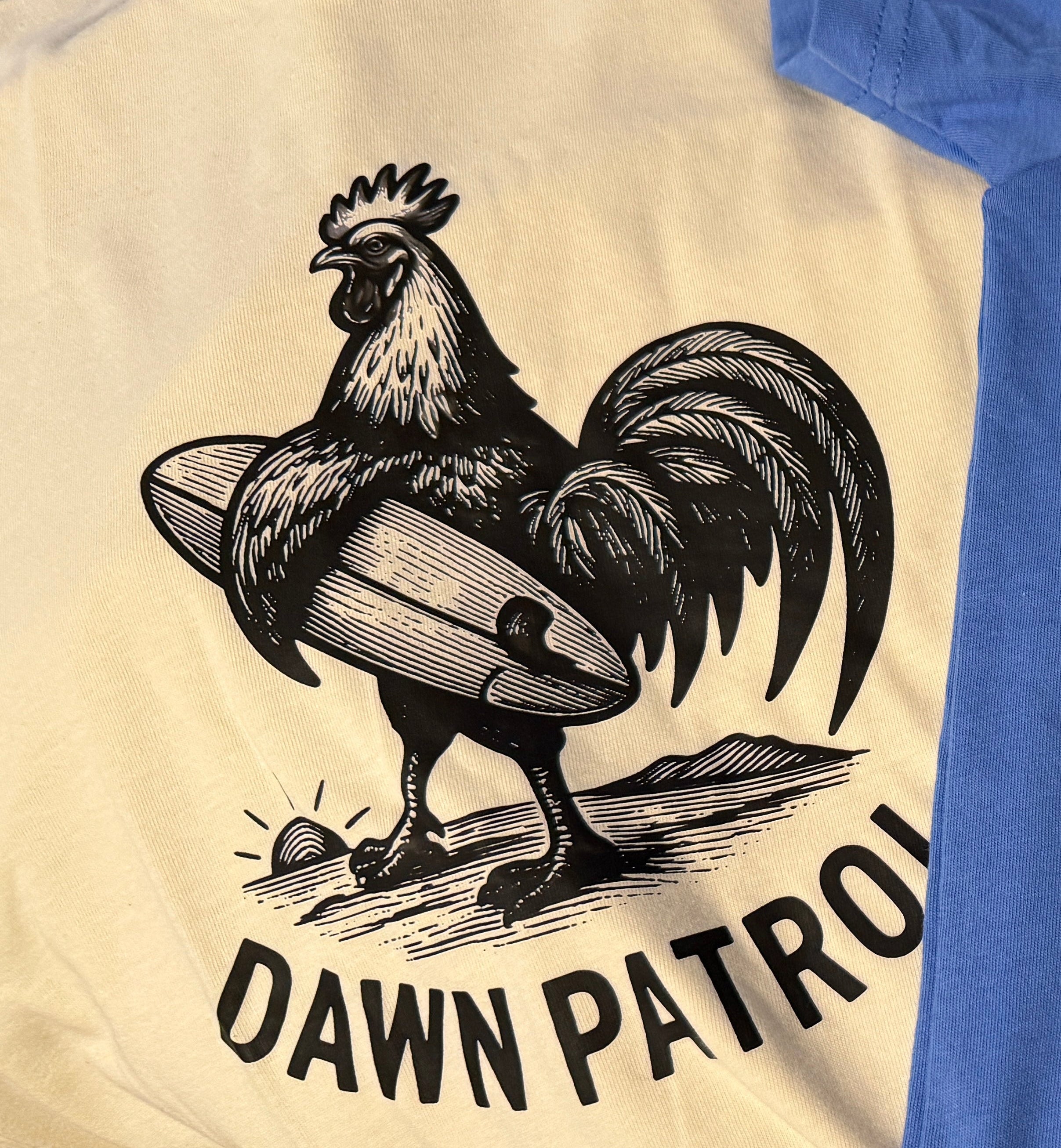 Two graphic t-shirts laid flat on a wooden floor. The top shirt is off-white with a black graphic of a rooster on a surfboard and the text 'DAWN PATROL'.