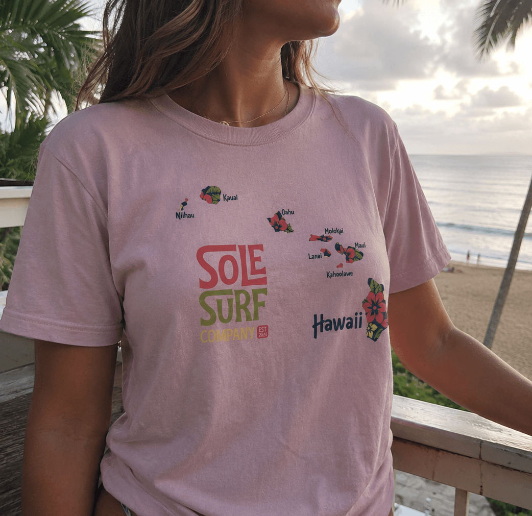 A woman wearing a light pink graphic t-shirt with 'SOLE SURF CHAMPION' and 'Hawaii' printed on the front, set against a beach background with palm trees.