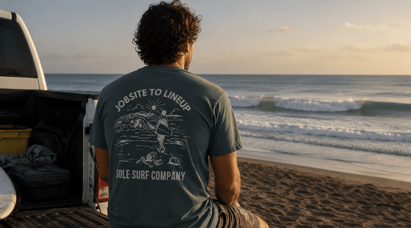 Man sitting on a truck bed at the beach wearing a t-shirt with a graphic design.