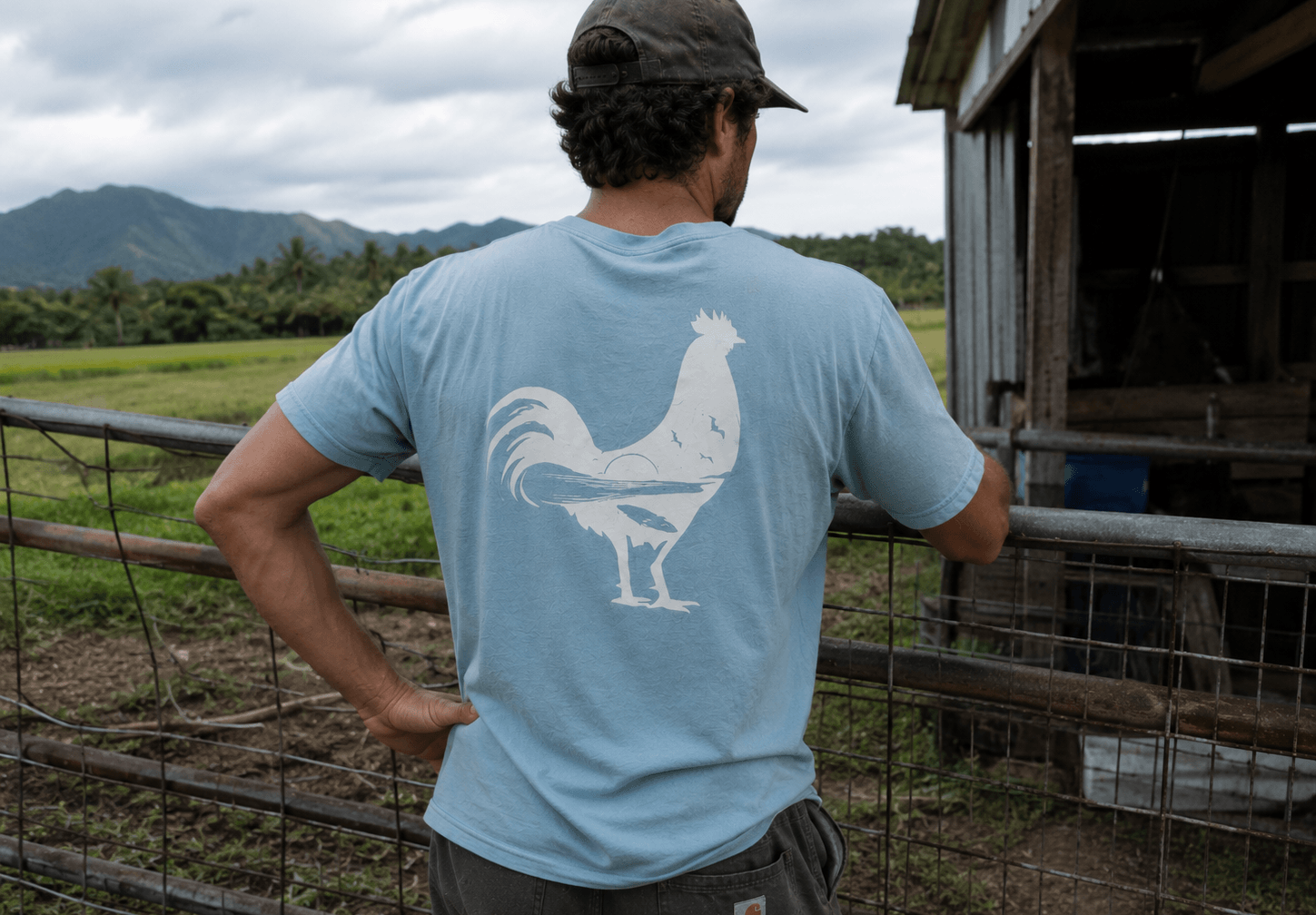 Person wearing a light blue t-shirt with a rooster design, standing in a farm setting.