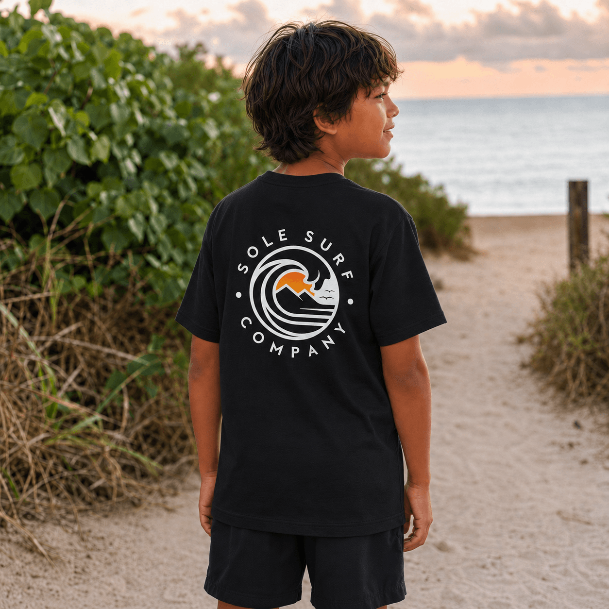 A child wearing a black t-shirt with a white and orange 'Sole Surf Company' logo on the back, standing on a sandy beach path with green plants and the ocean in the background.