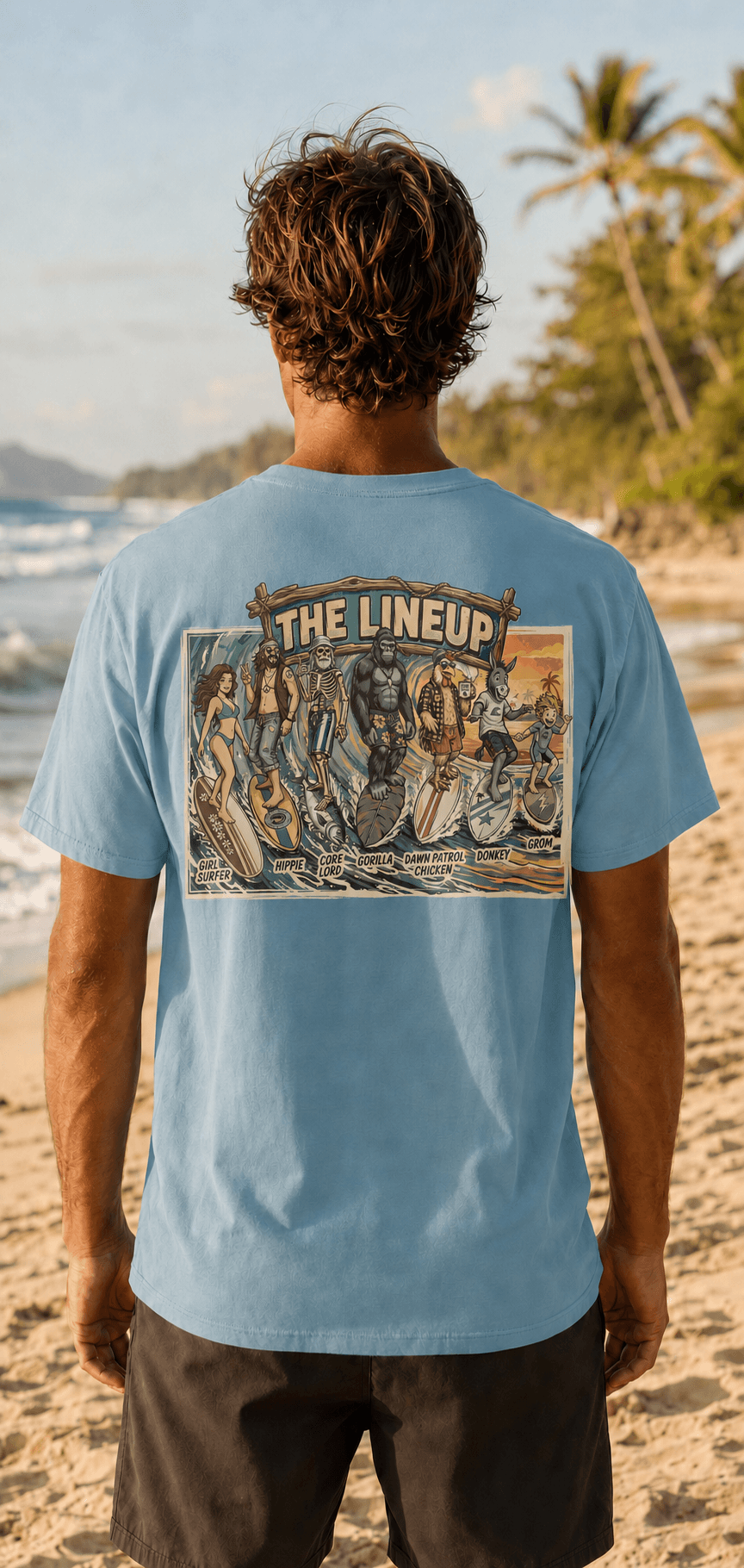 A man standing on a sandy beach with the ocean and palm trees in the background, viewed from behind. He is wearing a light blue t-shirt with a large graphic print on the back that reads 'THE LINEUP' and shows surfers, and dark shorts.