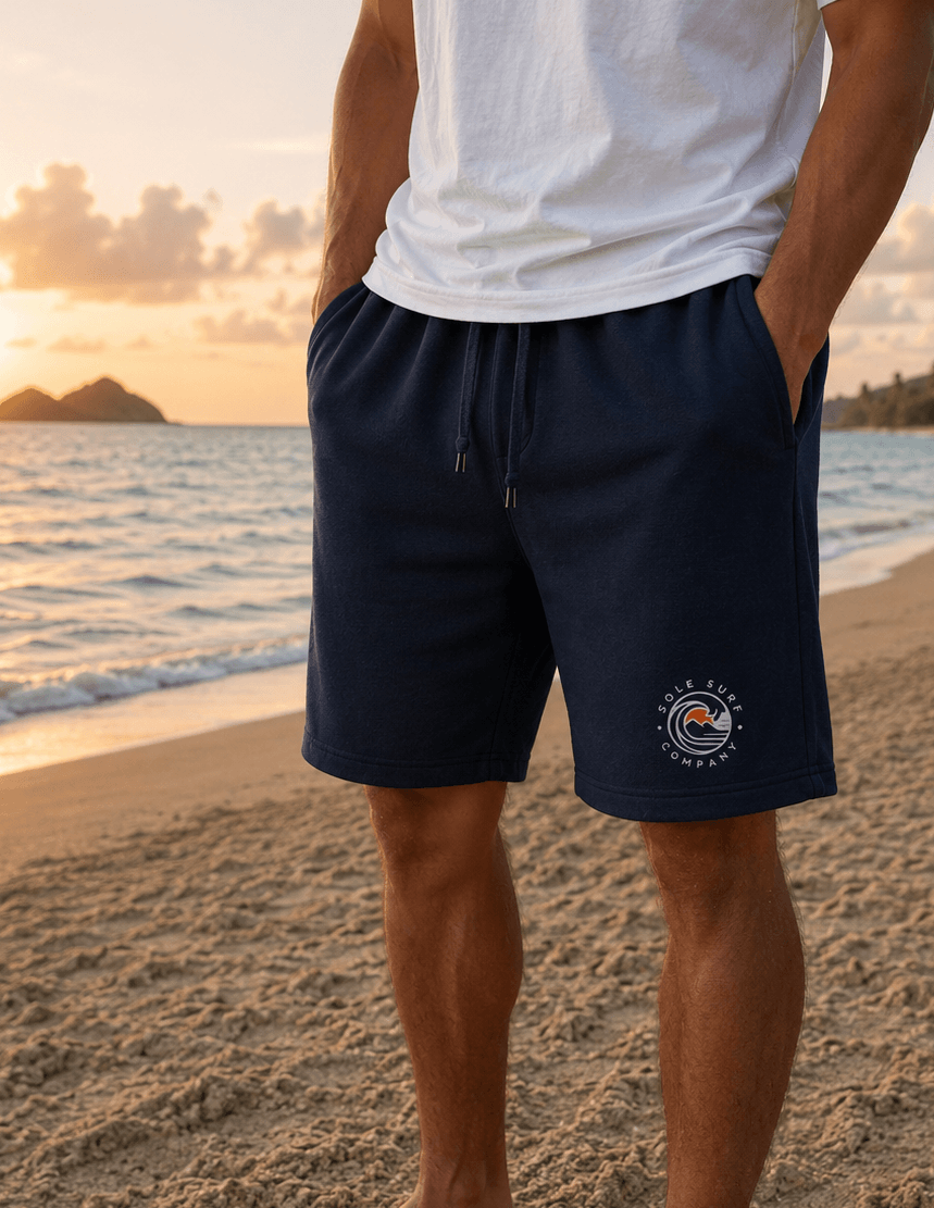 A man standing on a sandy beach at sunset wearing navy blue drawstring shorts with a circular 'The Life Coach' logo on the lower left leg, paired with a white t-shirt.
