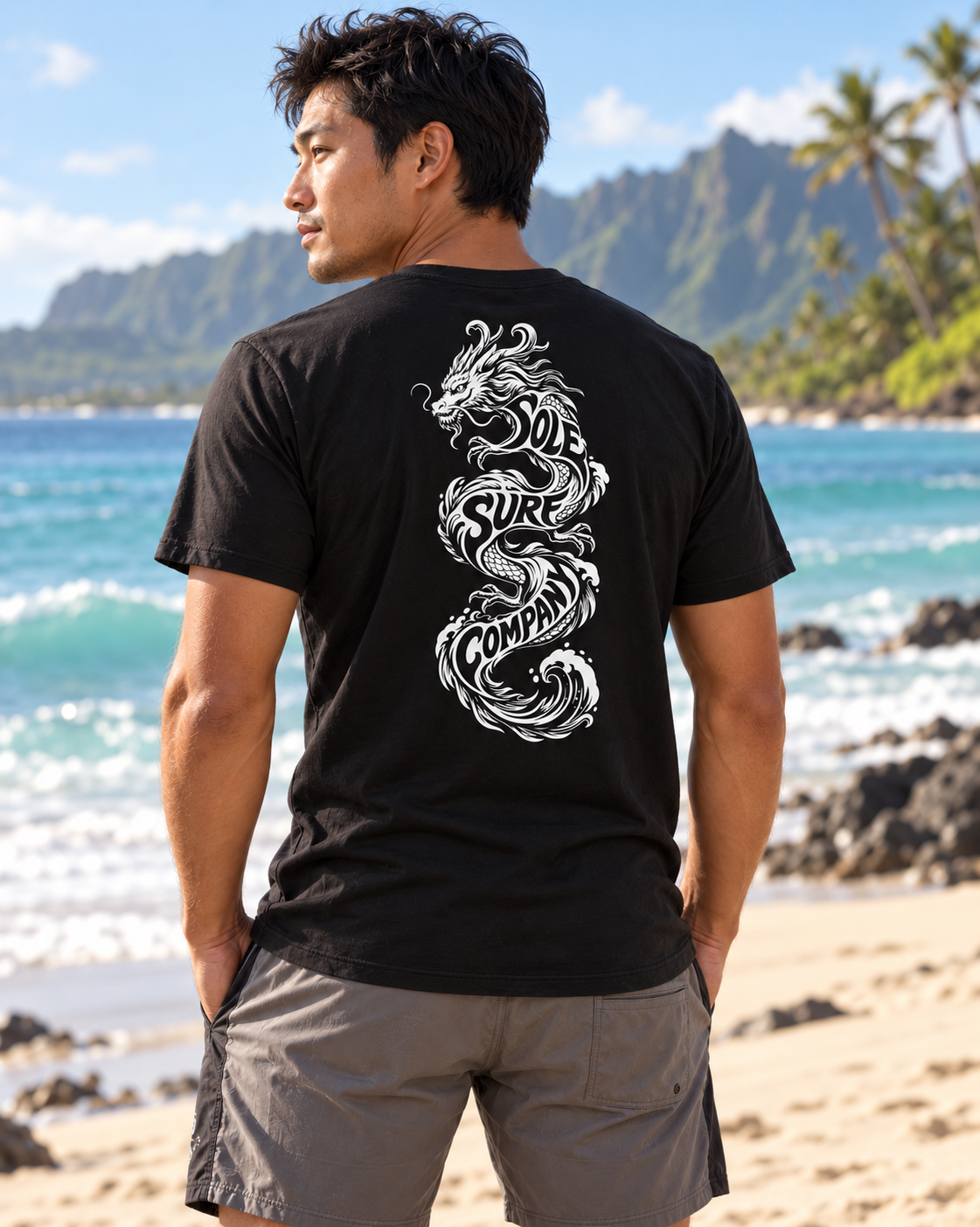 A man standing on a beach wearing a black t-shirt with a large white dragon graphic on the back, paired with gray shorts, with a tropical beach and mountains in the background.