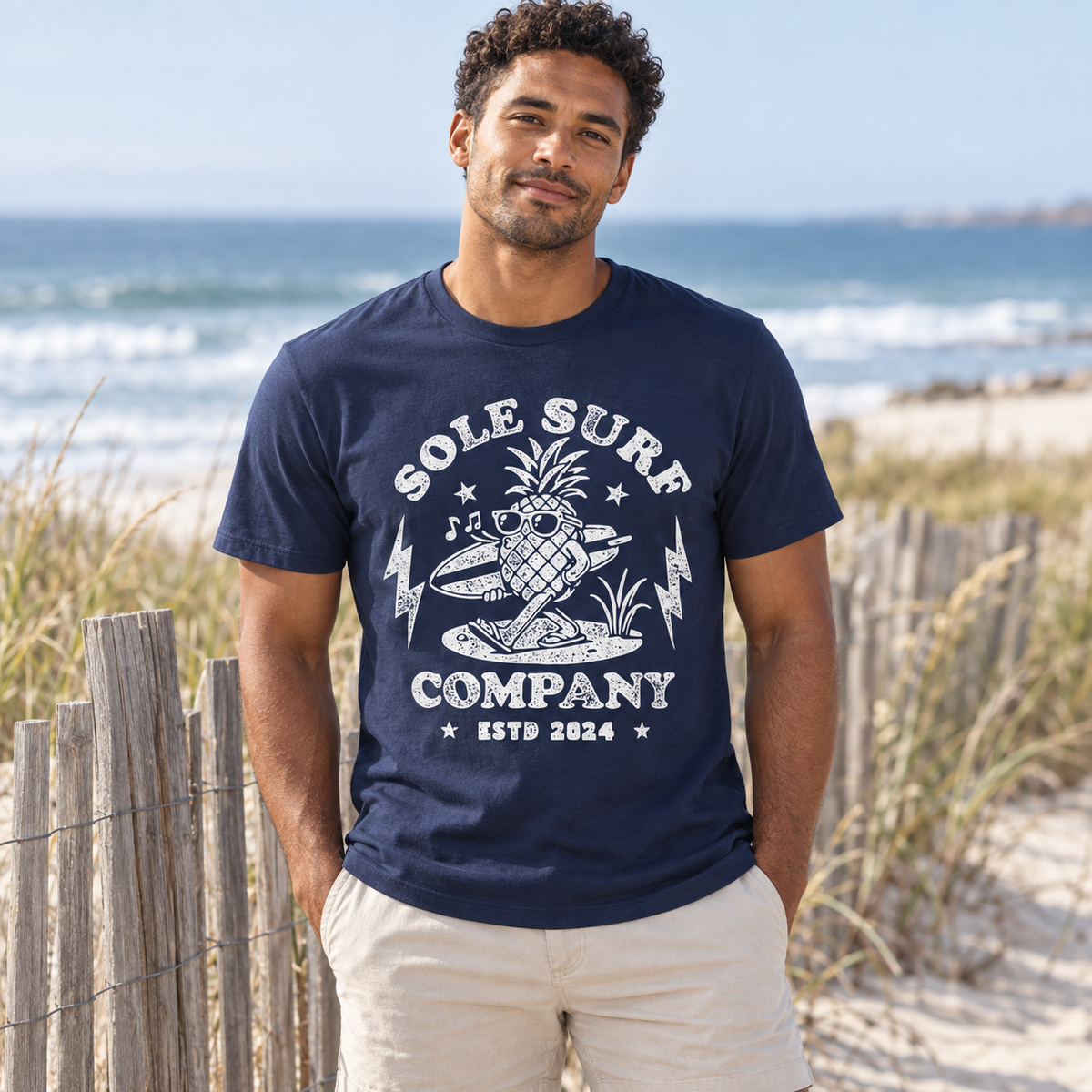 A man standing on a sandy beach wearing a navy blue graphic t-shirt with a white pineapple and surfboard design that reads 'SOLE SURF COMPANY ESTD 2024'.
