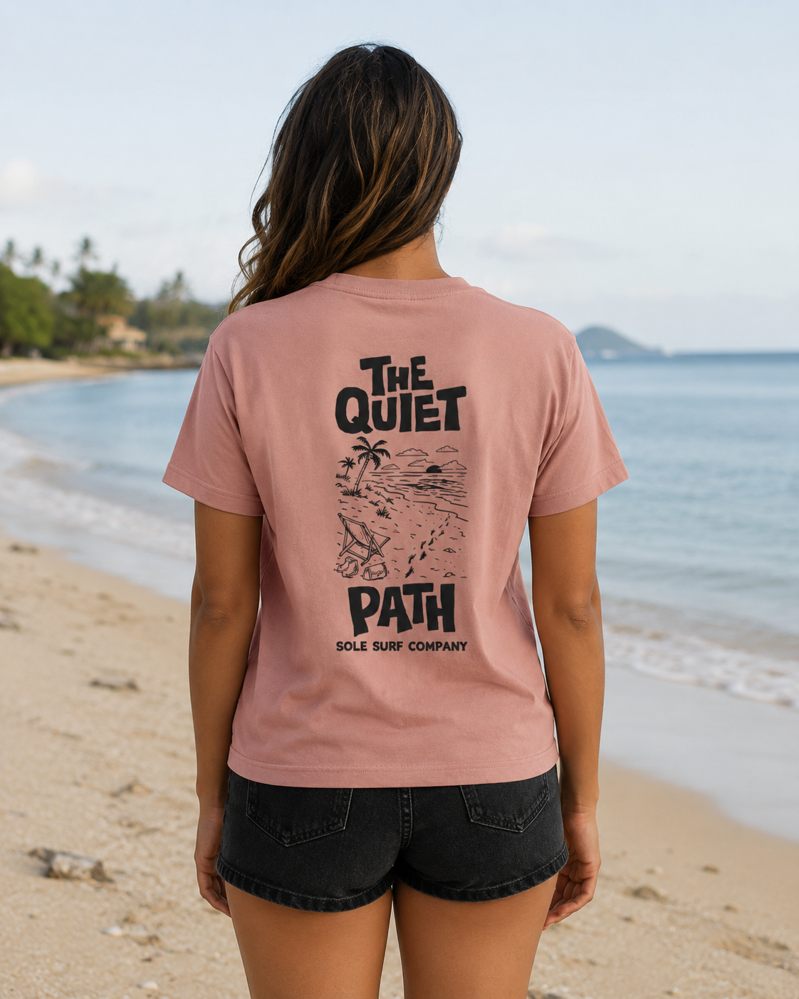 A woman stands on a sandy beach facing the ocean, wearing a dusty pink t-shirt with a black graphic print on the back that reads 'THE QUIET PATH SOLE SURF COMPANY' and black denim shorts.