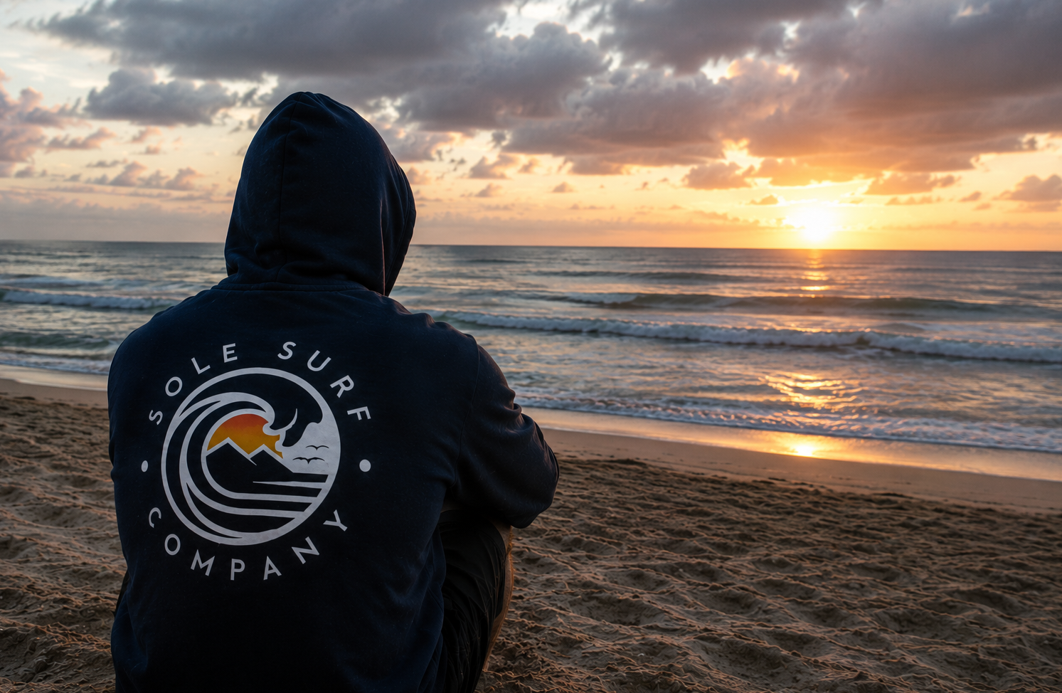 Person wearing a hoodie with 'Sole Surf Company' logo on a beach at sunset.