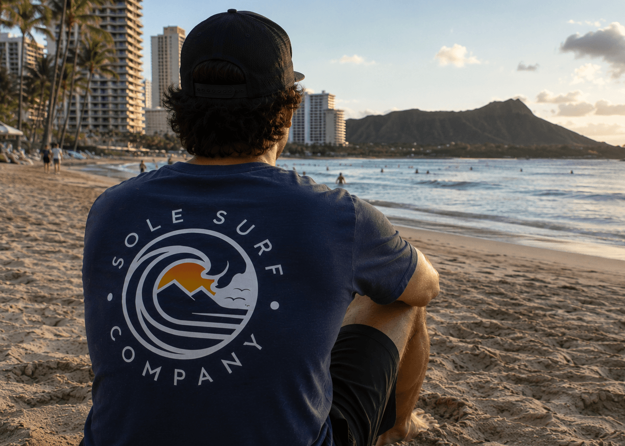 Person wearing a 'Sole Surf Company' t-shirt sitting on a beach with a scenic view of the ocean and mountains.