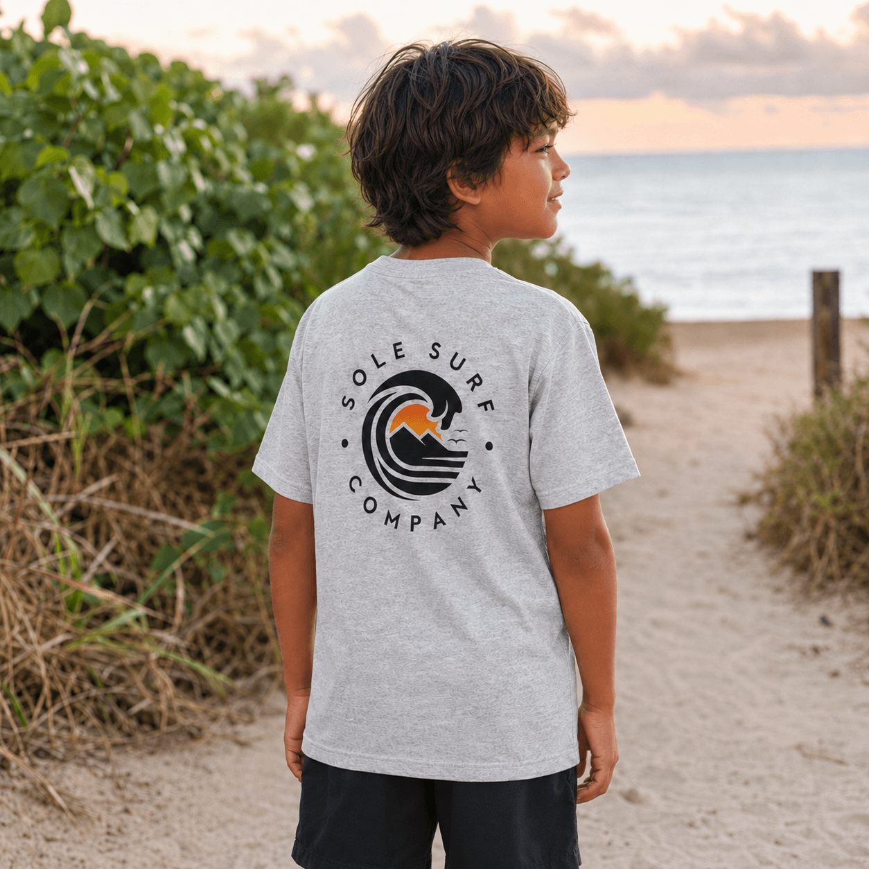 A child wearing a light gray heather t-shirt with a 'Sole Surf Company' wave graphic on the back, standing on a sandy beach path with green foliage and the ocean in the background.