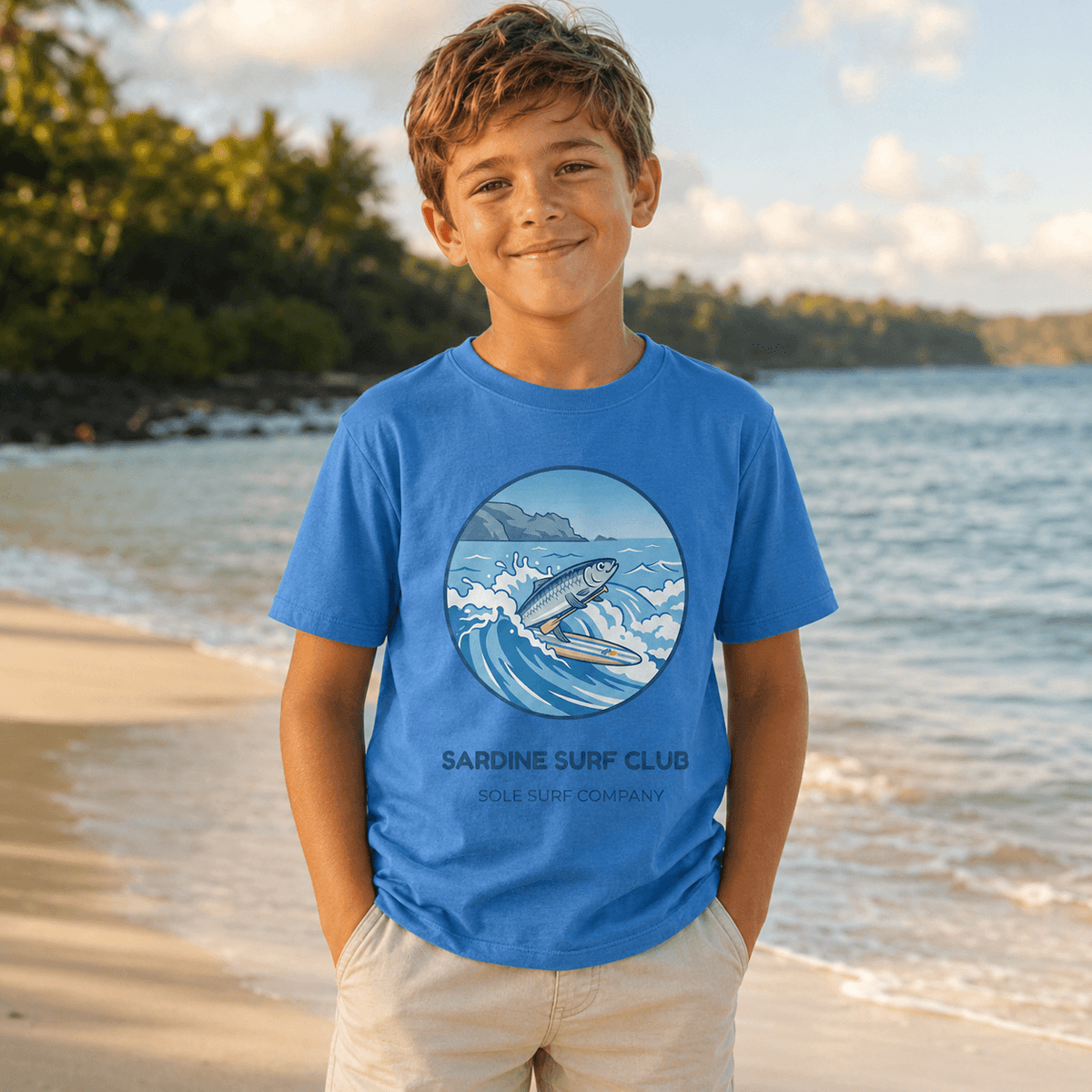 A young boy stands on a sandy beach wearing a blue t-shirt with a circular graphic of a surfer and the text 'SARDINE SURF CLUB'.