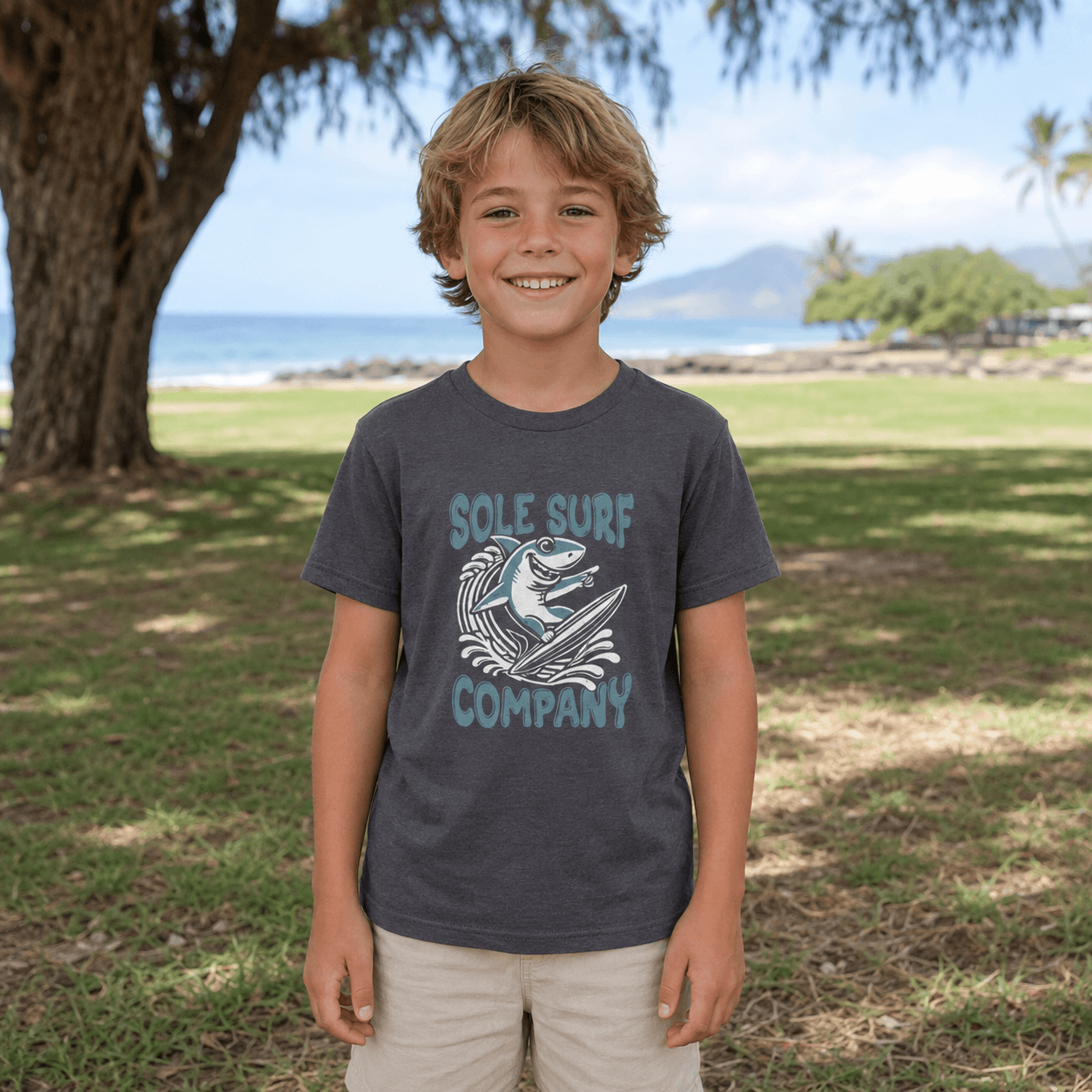 A smiling young boy stands outdoors on a grassy lawn near a tree, wearing a dark charcoal gray t-shirt with a light blue and white graphic that reads 'SOLE SURF COMPANY' and shows a shark and wave design. The background features a beach, ocean, and distant mountains under a bright sky.