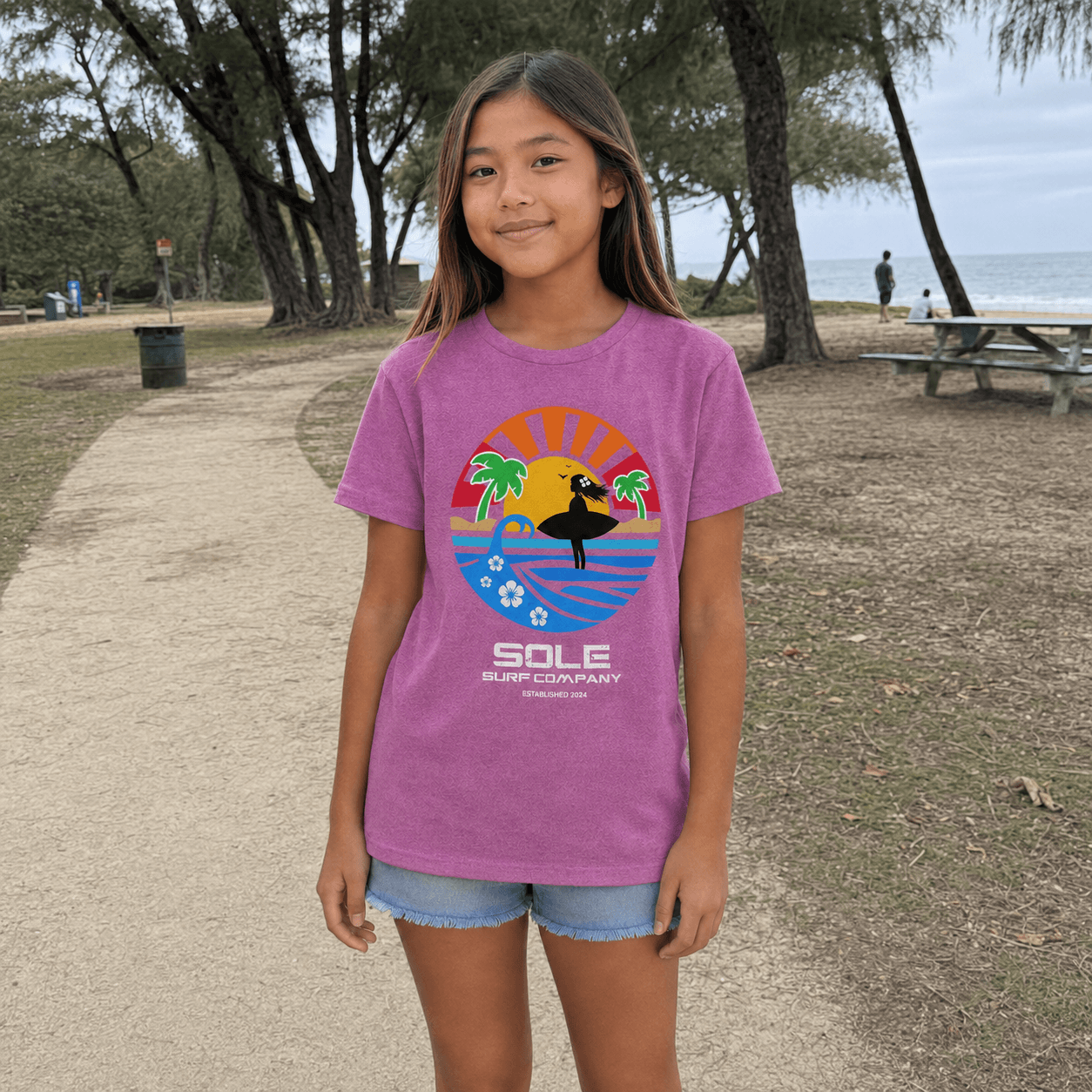 A young girl wearing a purple graphic t-shirt with a 'SOLE SURF COMPANY' design, standing on a sandy path in an outdoor park setting with trees and the ocean in the background.