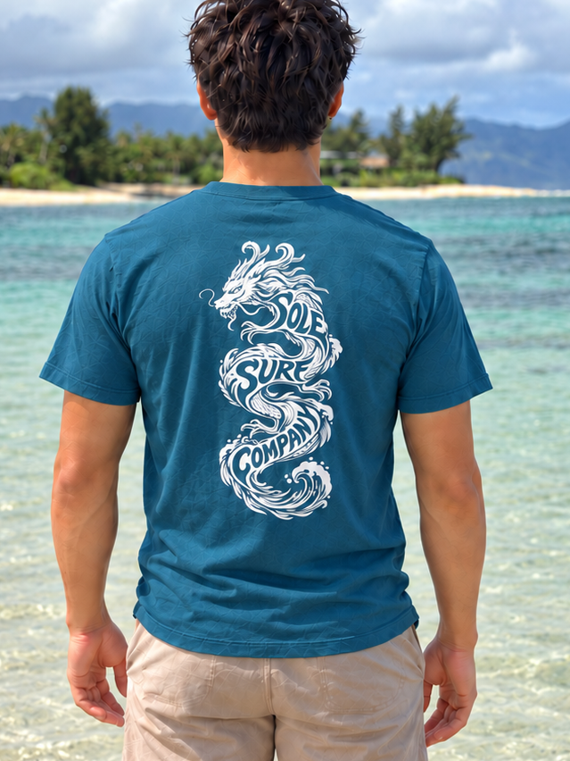 A man standing on a tropical beach, viewed from behind, wearing a teal blue t-shirt with a large white dragon graphic on the back and beige shorts.
