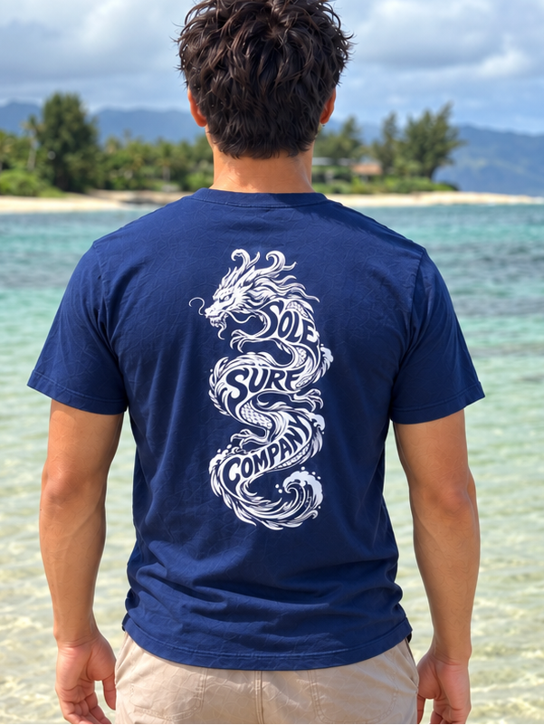 A man standing on a tropical beach, viewed from behind, wearing a navy blue t-shirt with a large white dragon graphic on the back and beige shorts.