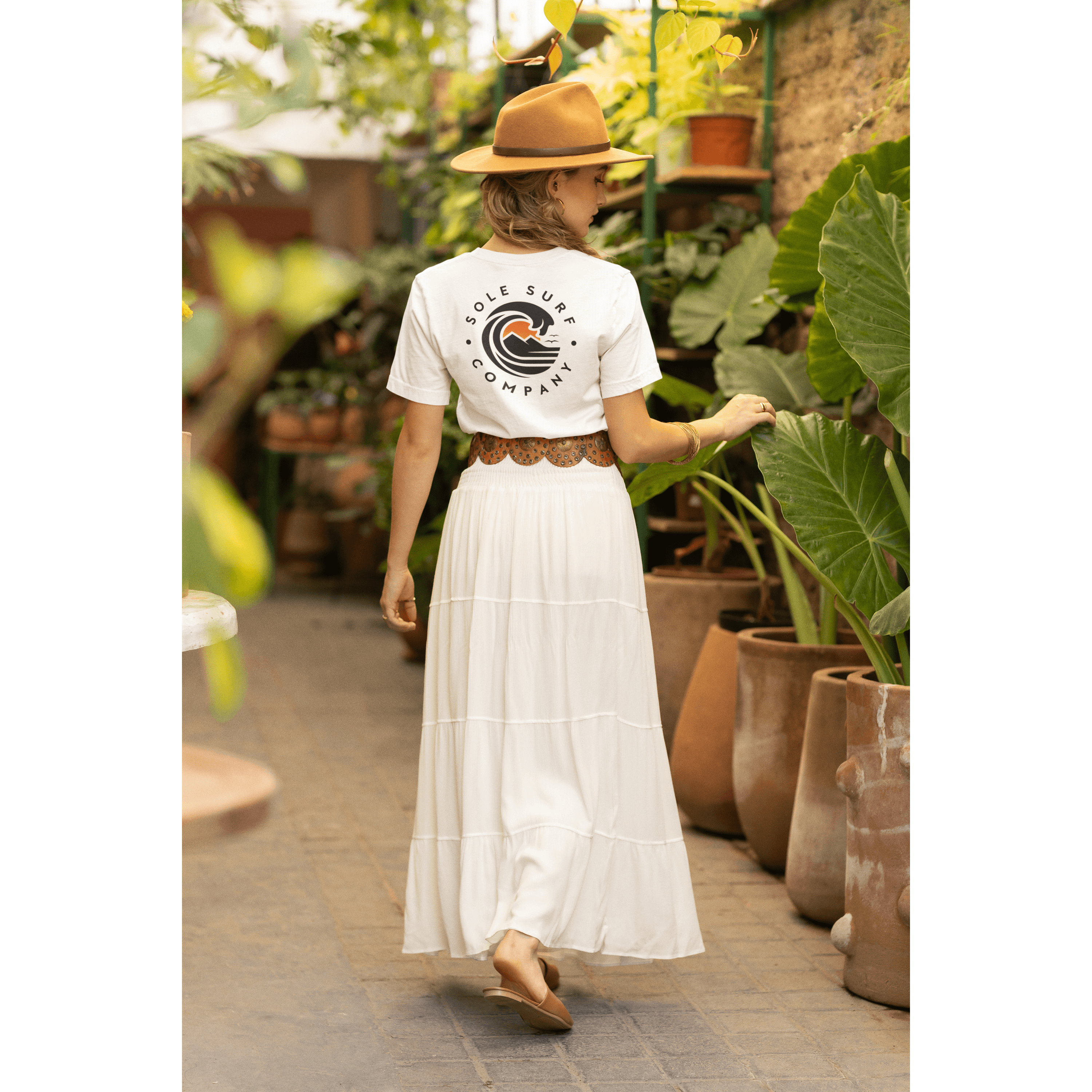 Woman wearing a white logo tee and a flowy skirt in a lush garden setting, showcasing a stylish, casual look.