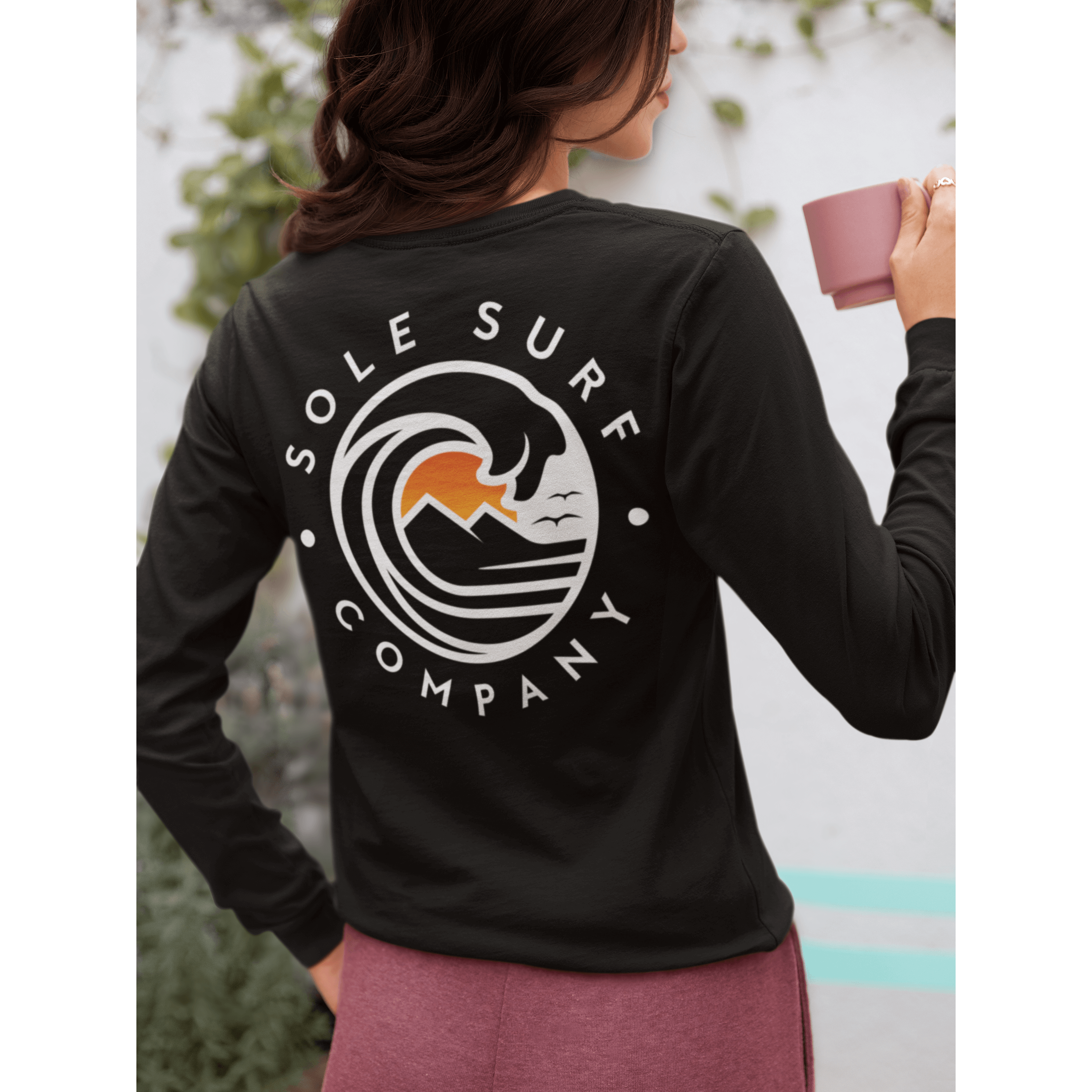 A woman wearing an SSC White Logo Long Sleeve surf tee, enjoying a drink with a coastal-themed design on the back.
