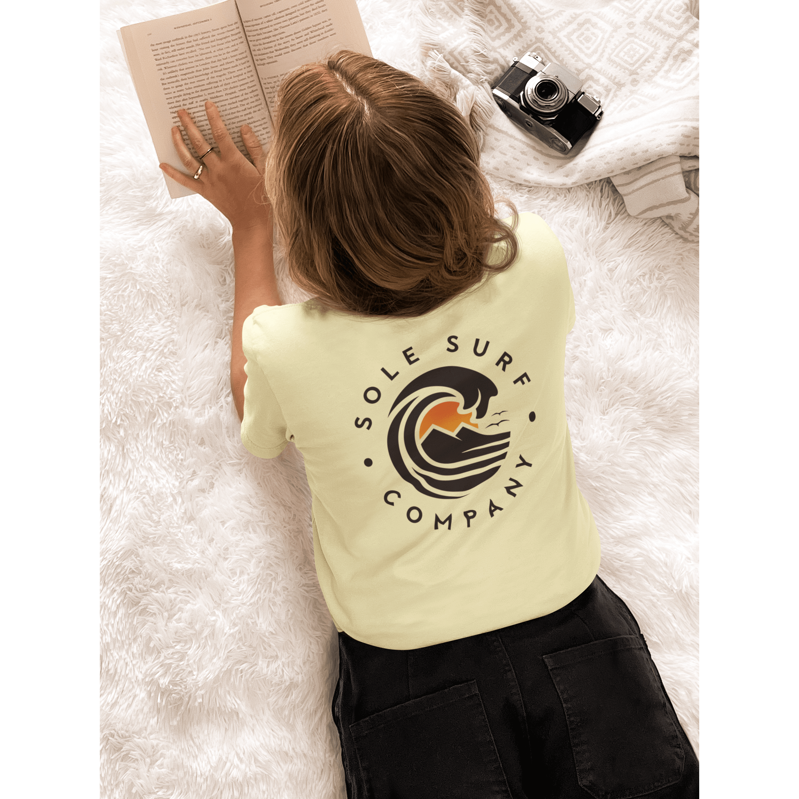 Woman wearing a light yellow tee with Sole Surf Company logo, reading a book on a cozy blanket.