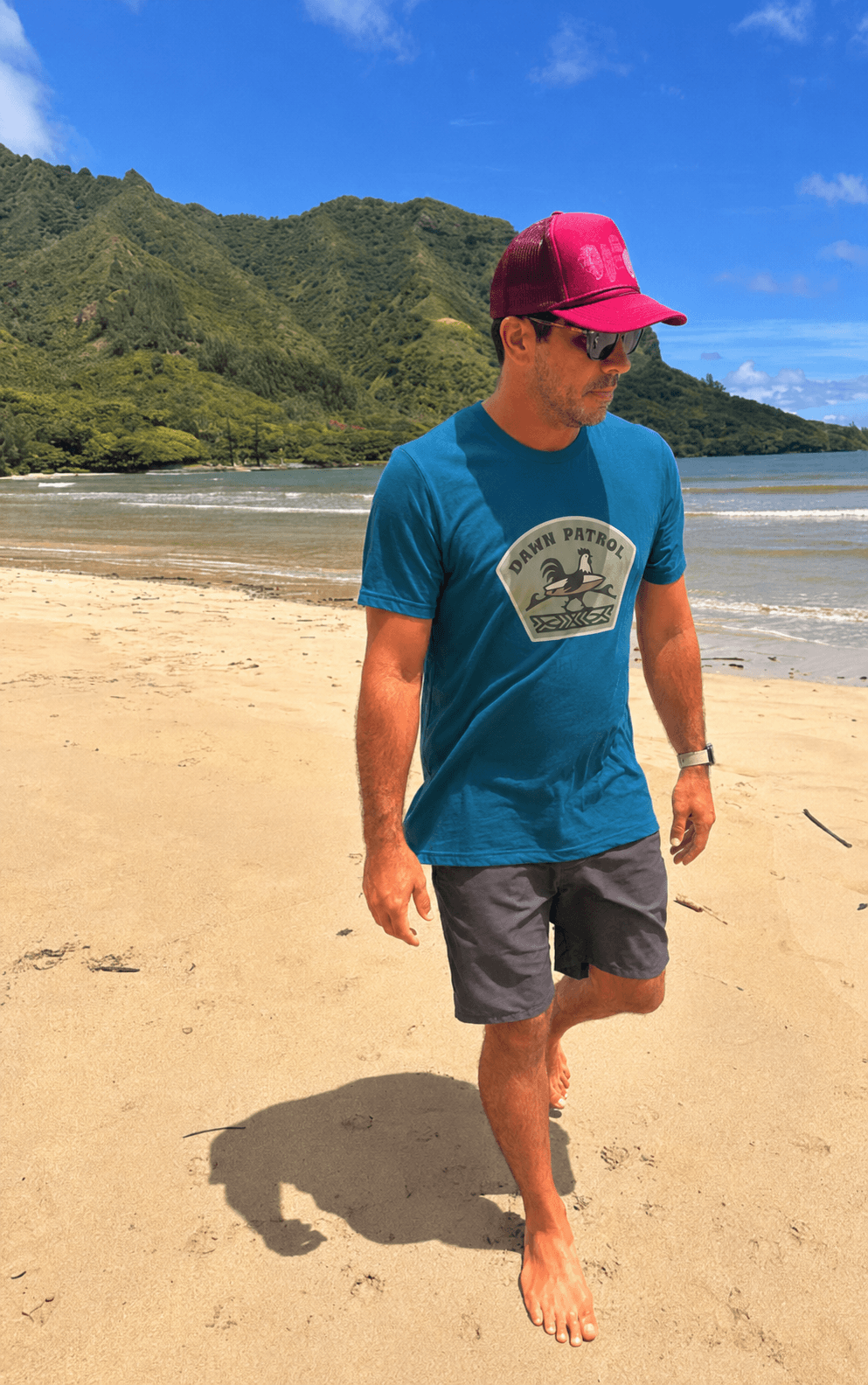 A man walking barefoot on a sandy beach wearing a blue graphic t-shirt, dark gray shorts, and a pink cap, with ocean waves and green mountains in the background.