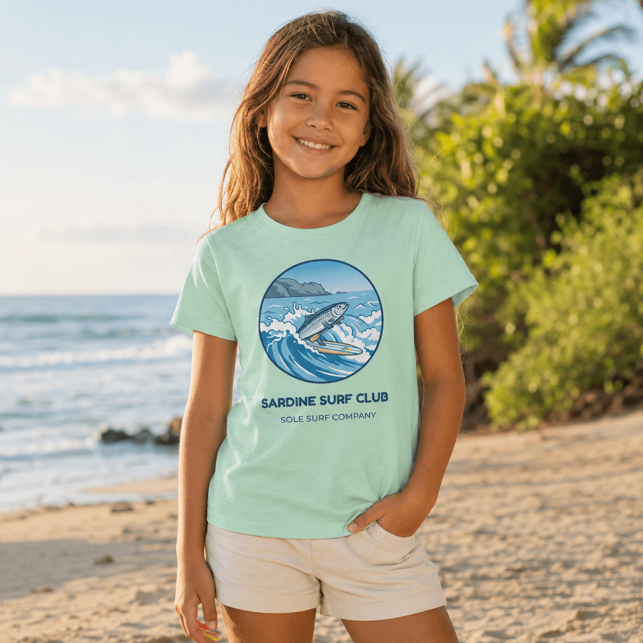 A young girl stands on a sandy beach wearing a mint green graphic t-shirt with a circular 'Sardine Surf Club' print and light beige shorts. The background features the ocean, sand, and green tropical foliage.