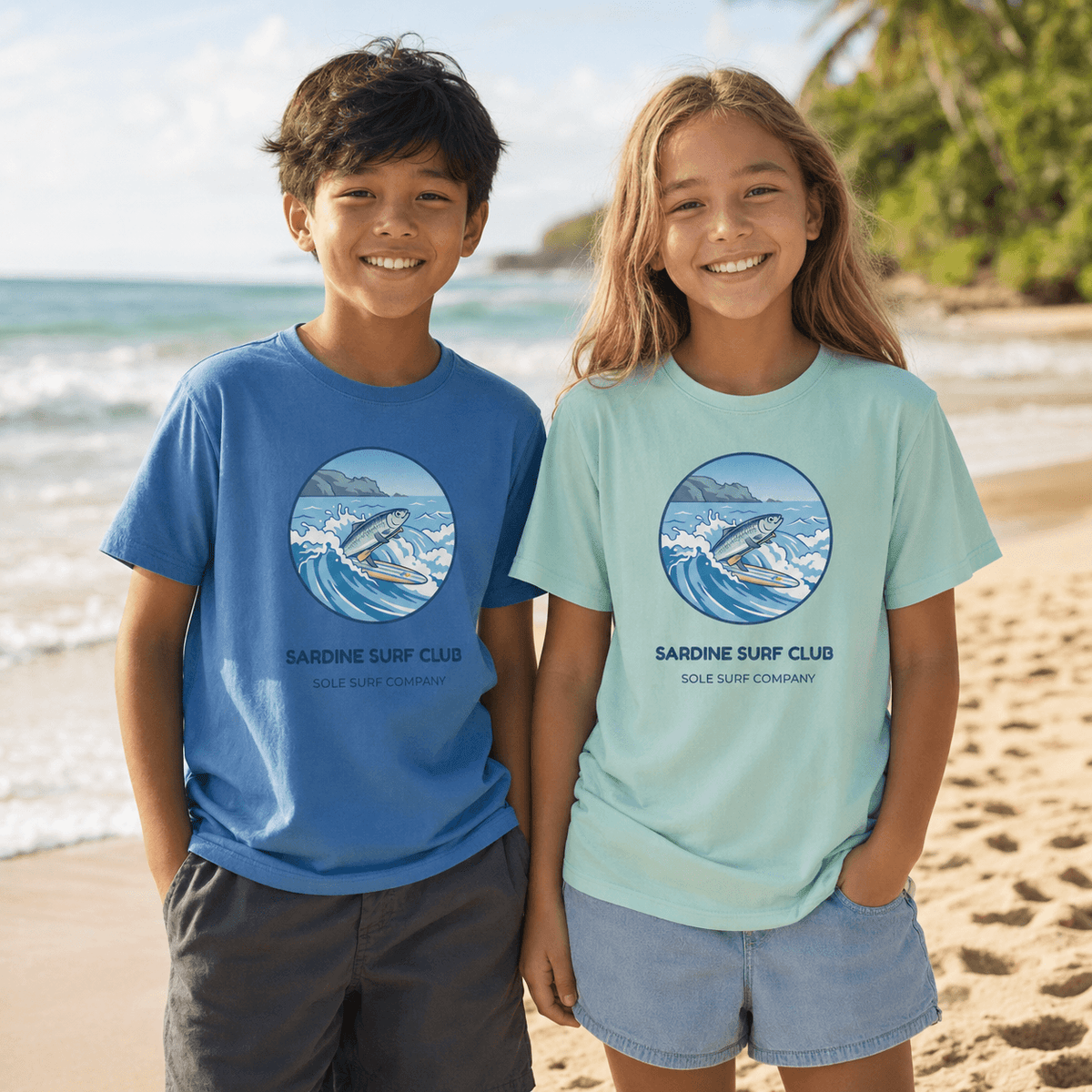 Two children standing on a sandy beach wearing matching graphic t-shirts with the 'Sardine Surf Club' logo. The boy wears a medium blue t-shirt, and the girl wears a mint green t-shirt.