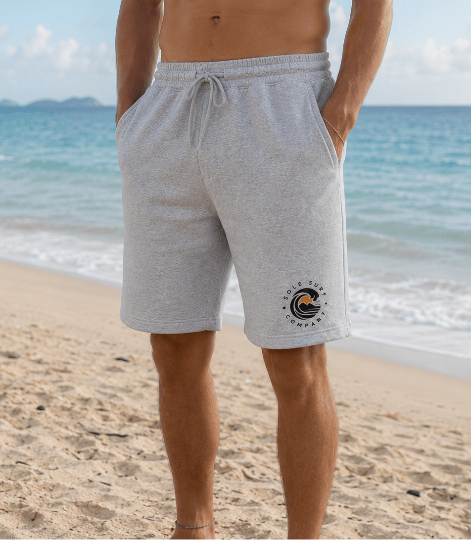 A man standing on a sandy beach wearing light heather gray drawstring shorts with a circular 'The Beach Company' logo printed on the lower left leg. The background shows the blue ocean and sky.