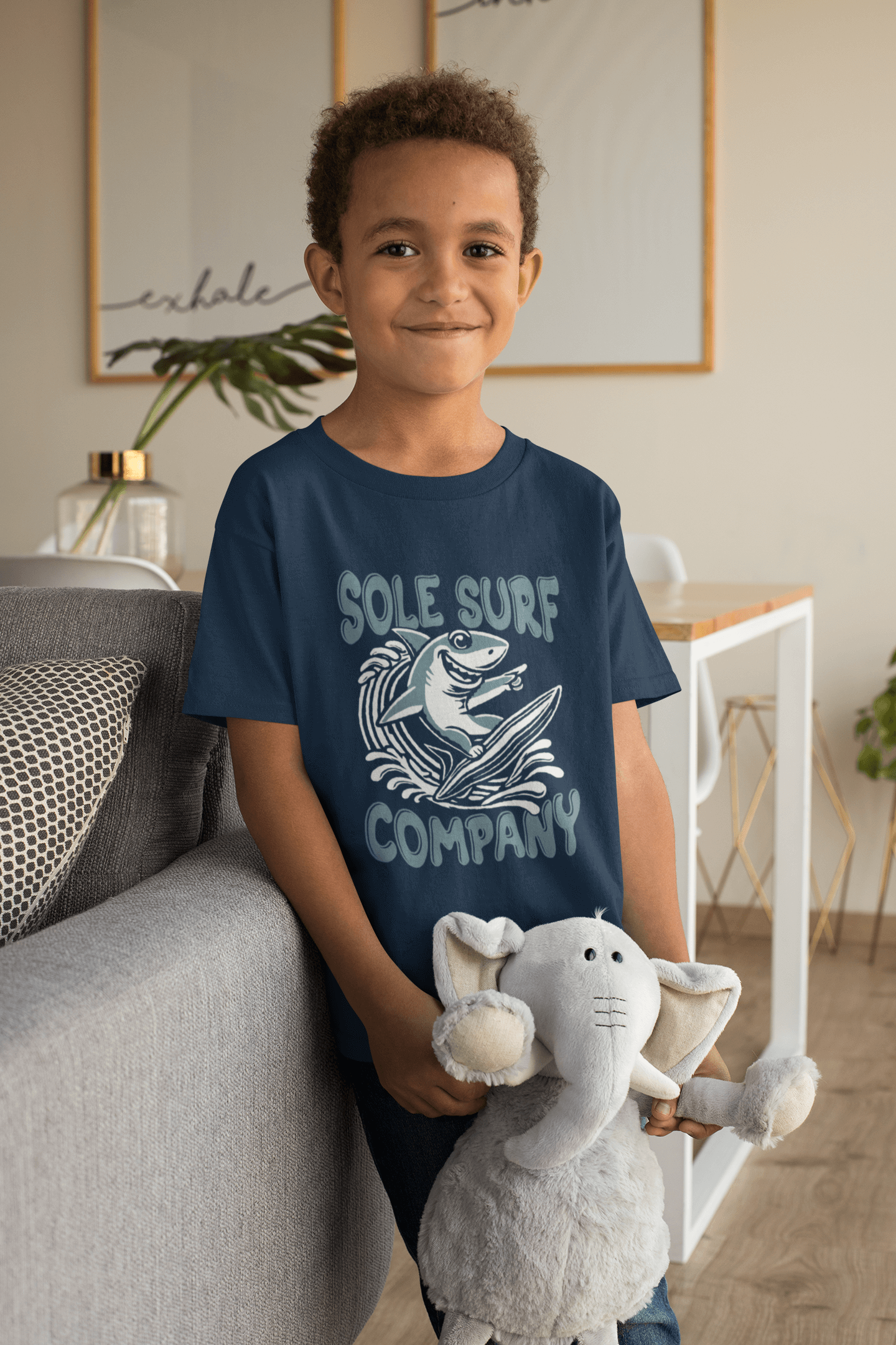 Smiling boy in navy blue Kids Surfing Shark Tee holding a plush elephant, showcasing playful ocean-themed design.