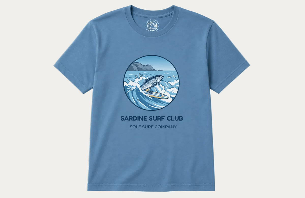 A light blue short-sleeve crewneck t-shirt featuring a circular graphic of a surfer riding a wave, with the text 'SARDINE SURF CLUB' and 'SOLE SURF COMPANY' printed below it, displayed on a clean off-white background.