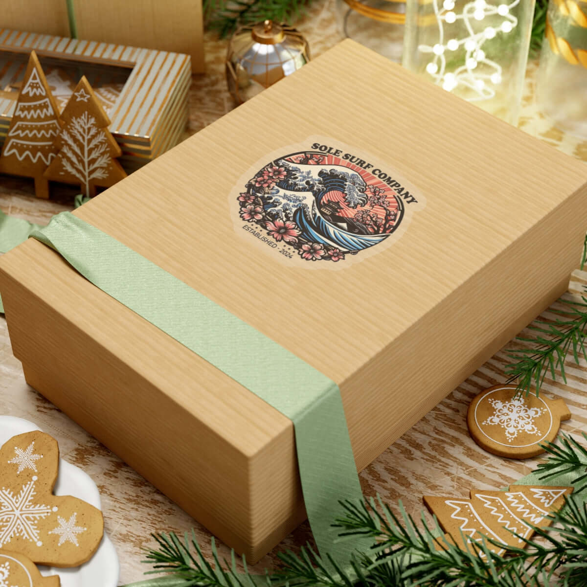 Gift box with logo featuring a wave design, decorated with a green ribbon and surrounded by holiday decor.