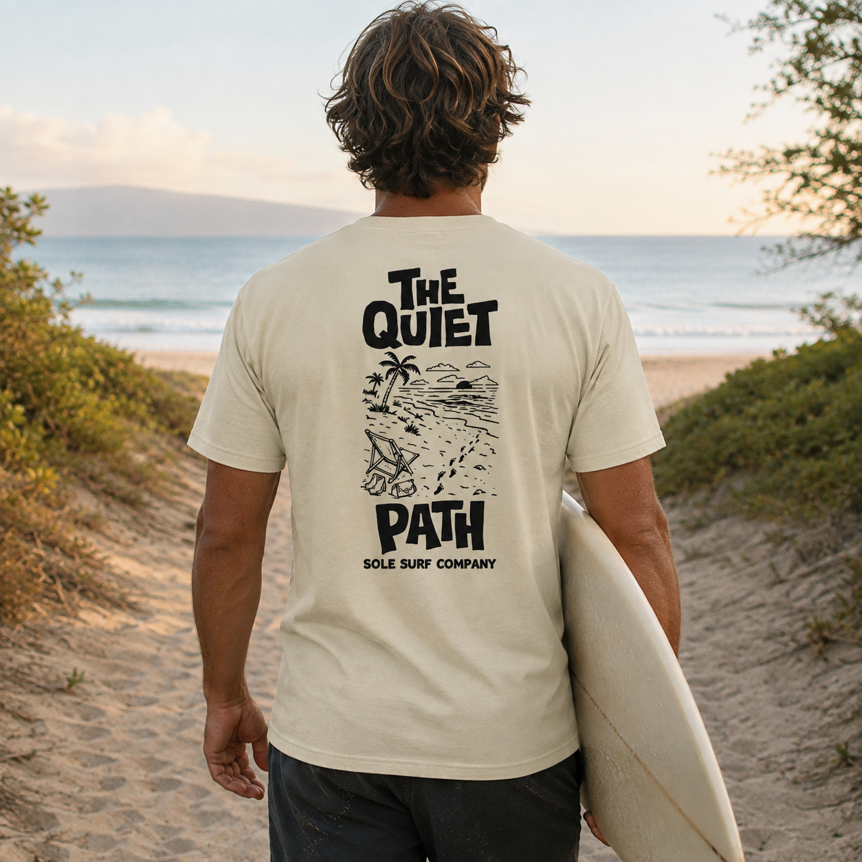 A man on a beach, seen from behind, wearing a cream-colored t-shirt with a black graphic print that reads 'THE QUIET PATH SOLE SURF COMPANY' and holding a white surfboard.