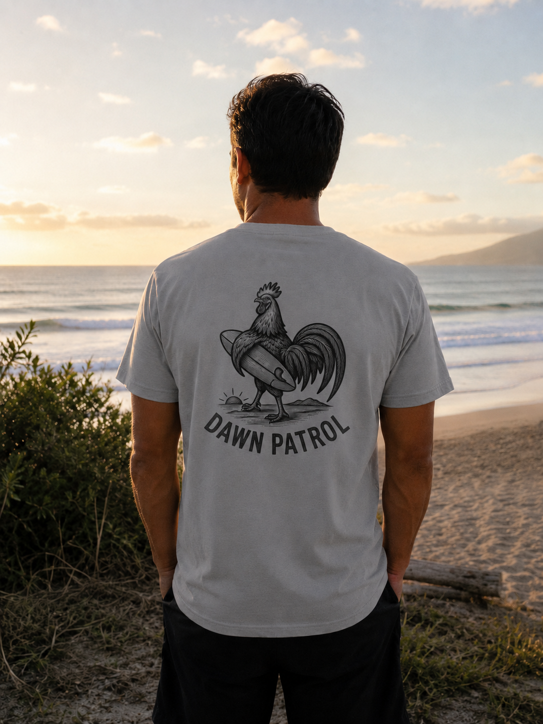 A man standing on a beach at sunset, viewed from behind, wearing a light gray t-shirt with a large black graphic of a rooster on a surfboard and the text 'DAWN PATROL' printed on the back.