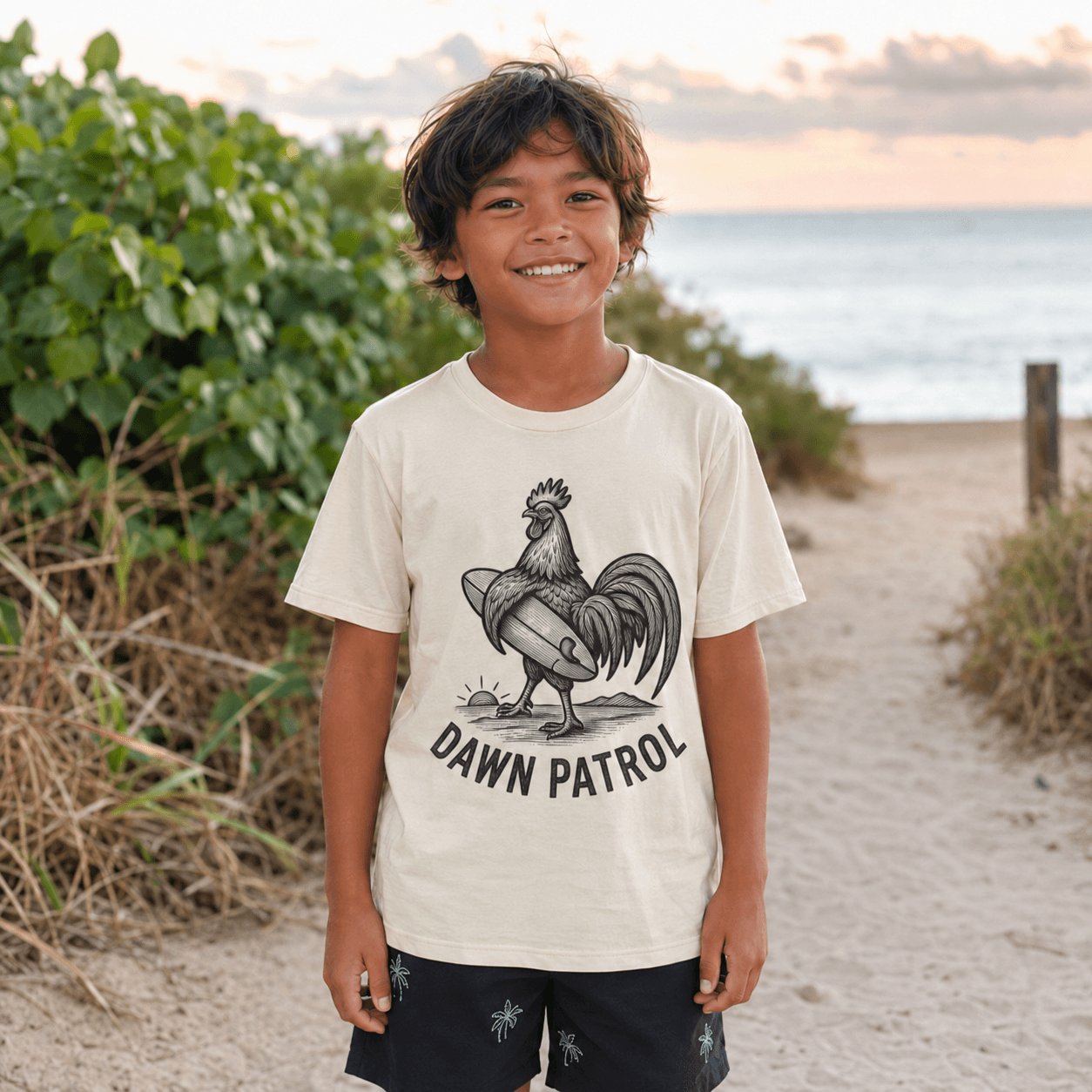 A smiling young boy stands on a sandy beach path wearing a cream-colored t-shirt with a black graphic of a rooster carrying a surfboard and the text 'DAWN PATROL'.