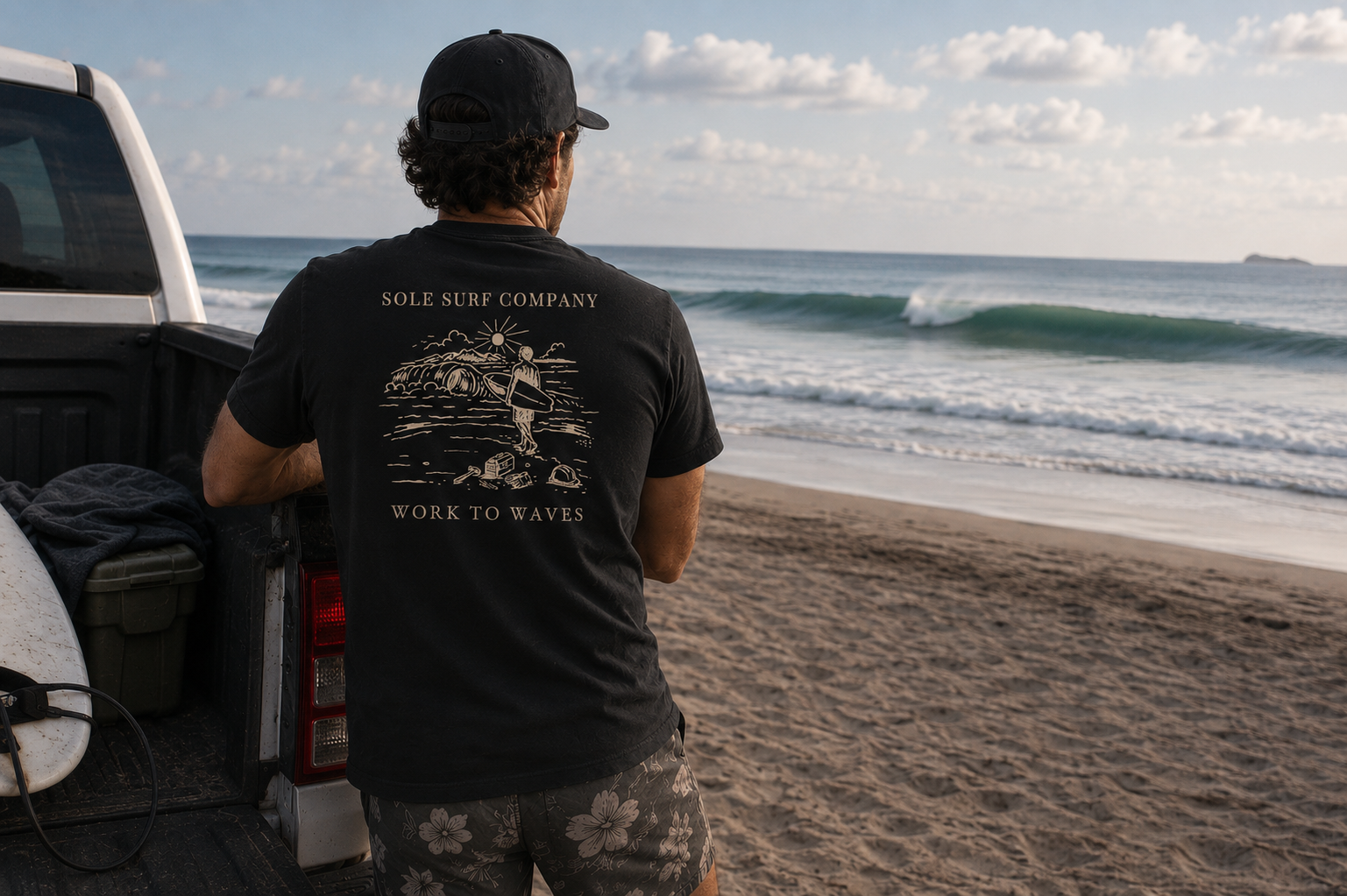 A man stands in the bed of a white pickup truck on a beach, facing the ocean. He is wearing a black t-shirt with a beige graphic print that reads 'SOLE SURF COMPANY' and 'WORK TO WAVES', along with a black cap and patterned shorts.