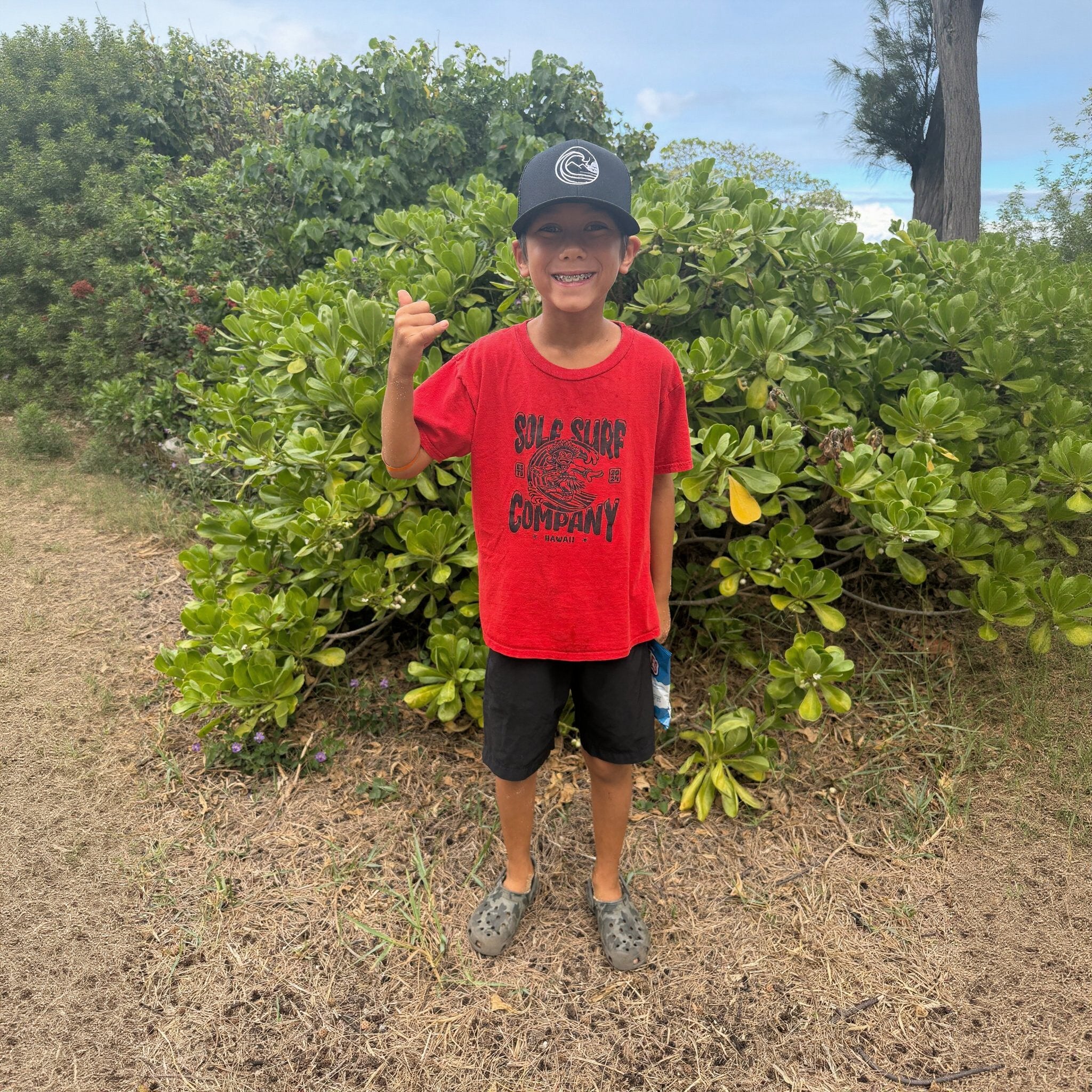 Child wearing SSC Trucker Cap and red Sole Surf Company t-shirt, standing in front of lush greenery.