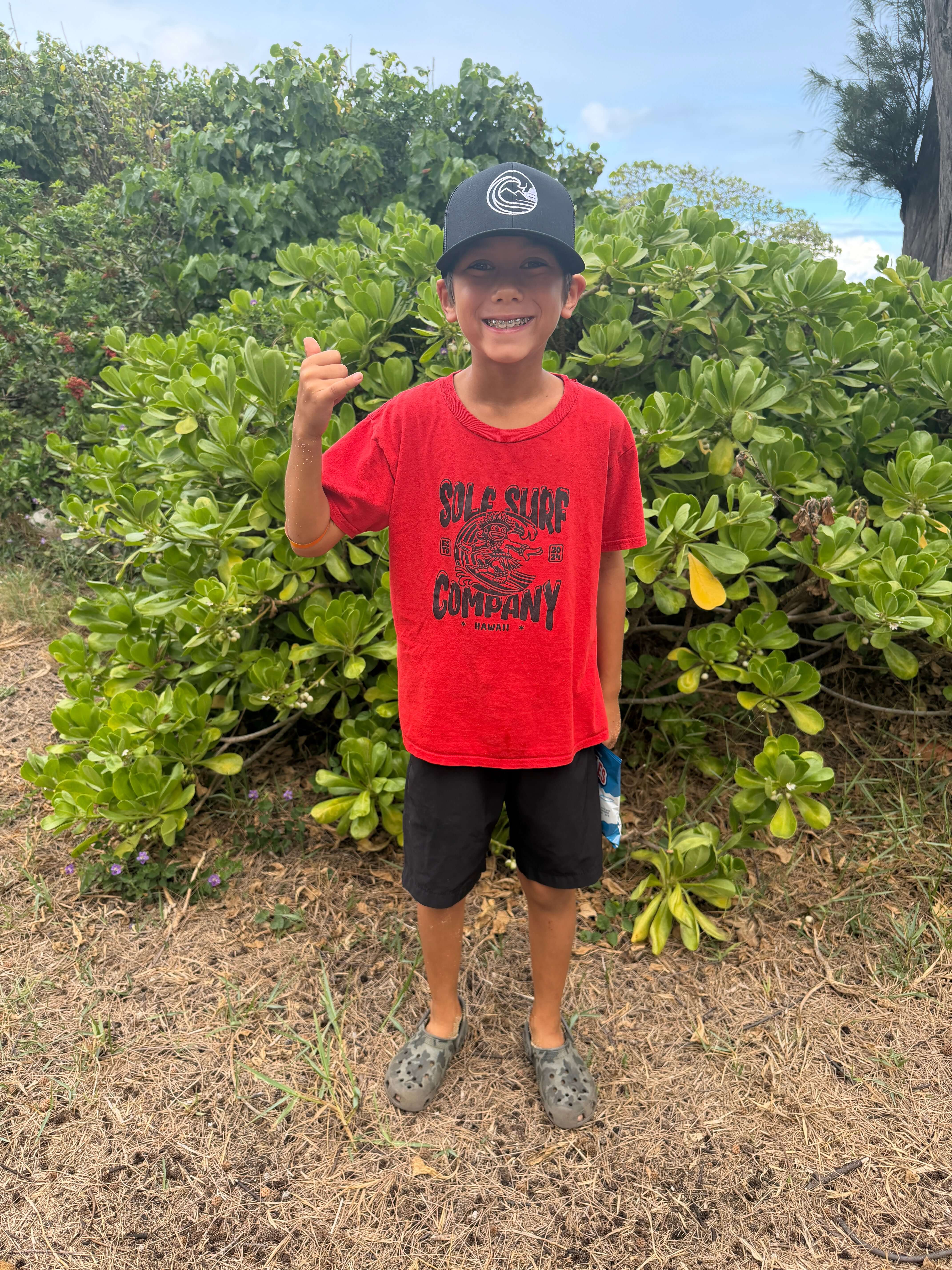 Child wearing SSC Trucker Cap and red Sole Surf Company t-shirt, standing in front of lush greenery.