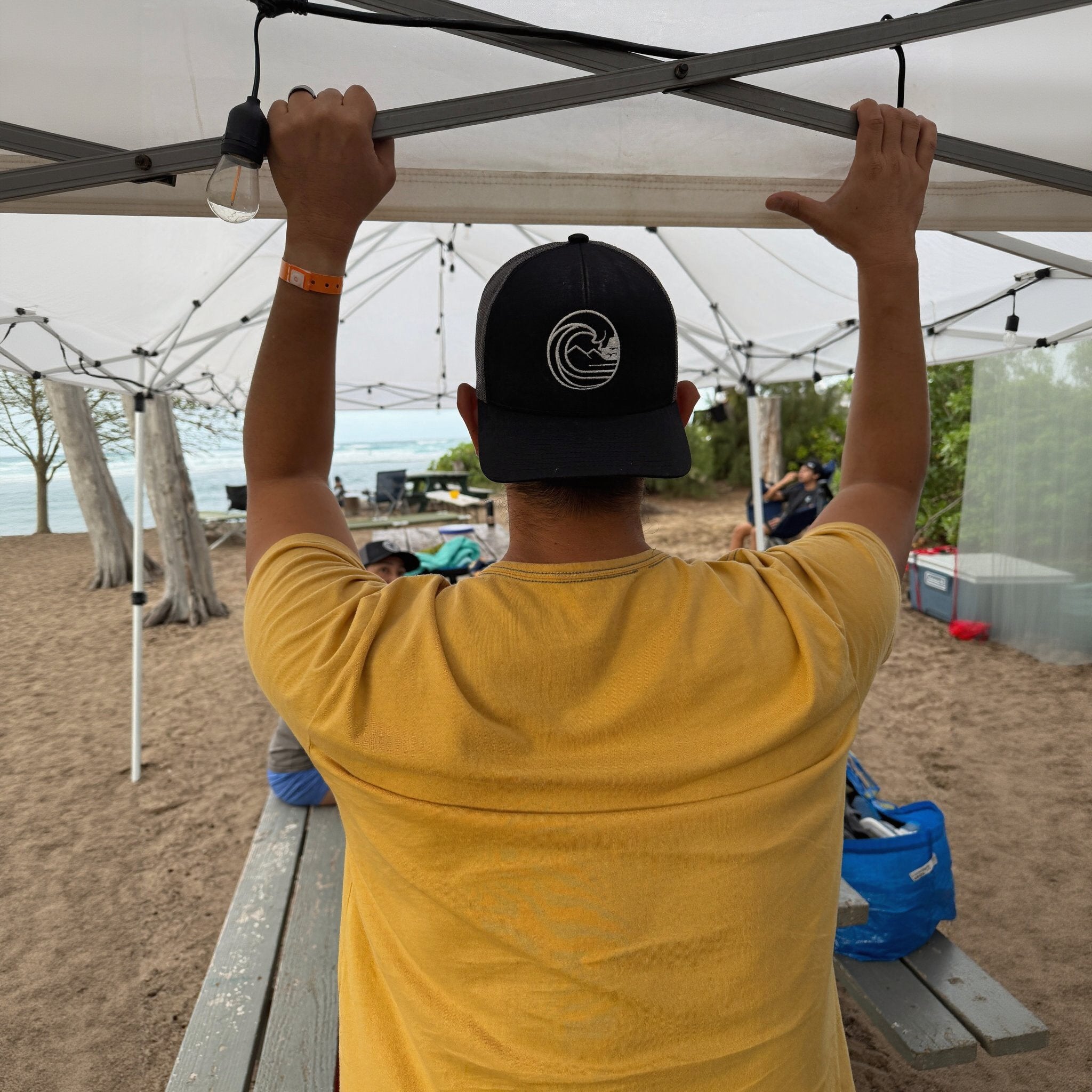 Person wearing SSC Trucker Cap, enjoying a beach day under a tent with friends.