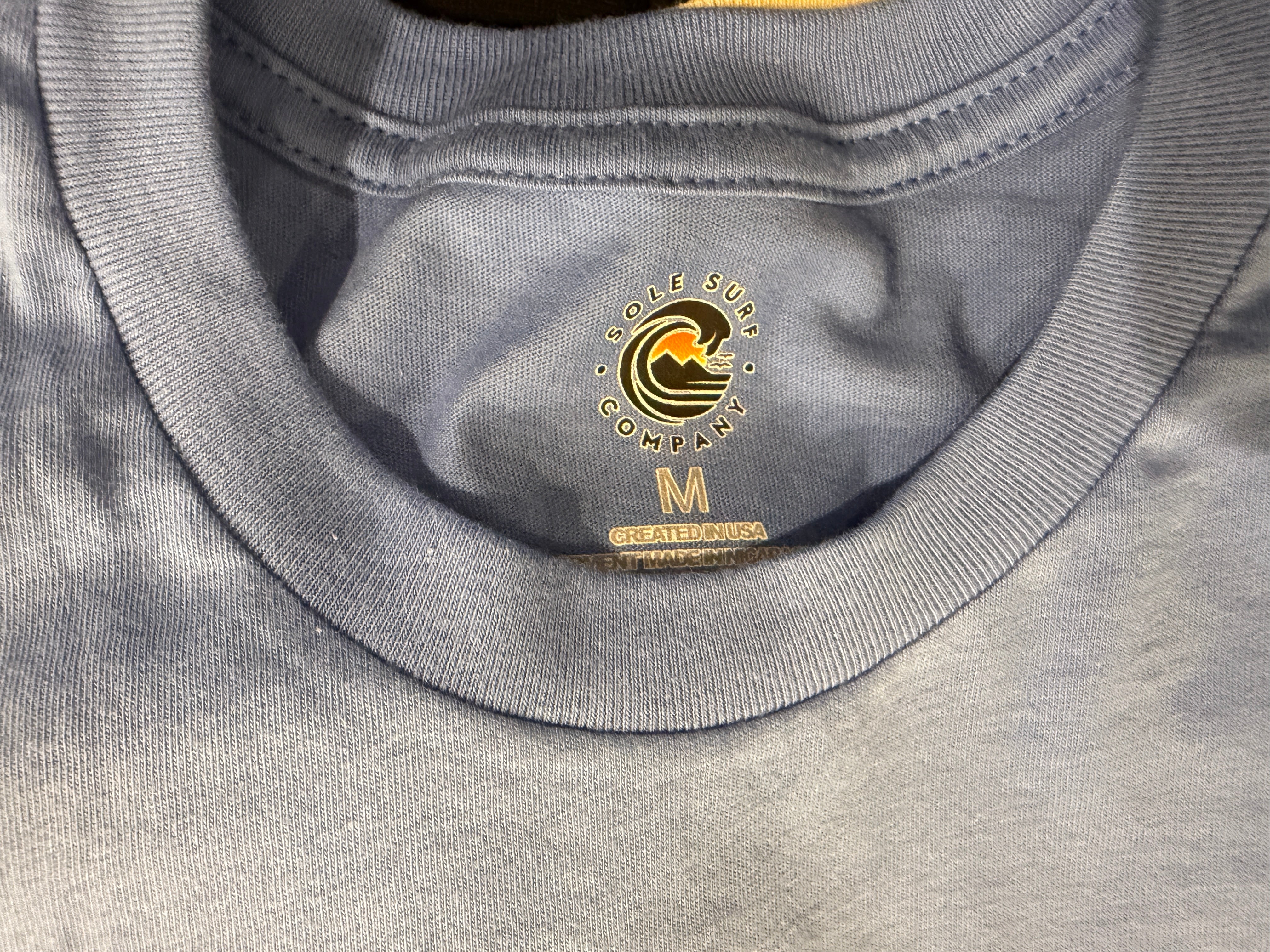 Close-up of a light blue-gray crewneck t-shirt showing the inner neck label with a circular logo that reads 'MINDSET COACHING' and 'M' size.