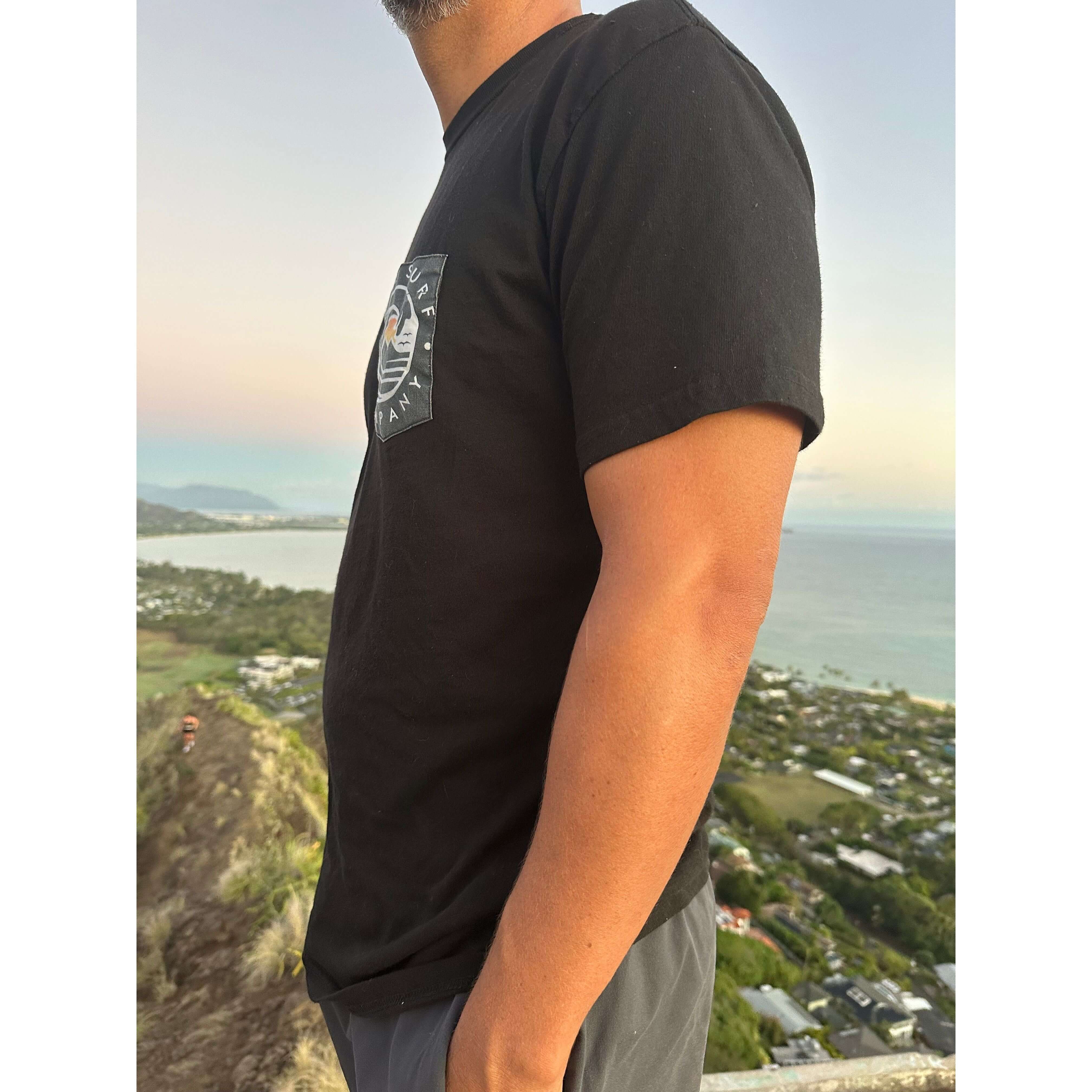 Side view of a man wearing a black logo pocket t-shirt against a scenic backdrop, ideal for casual wear.